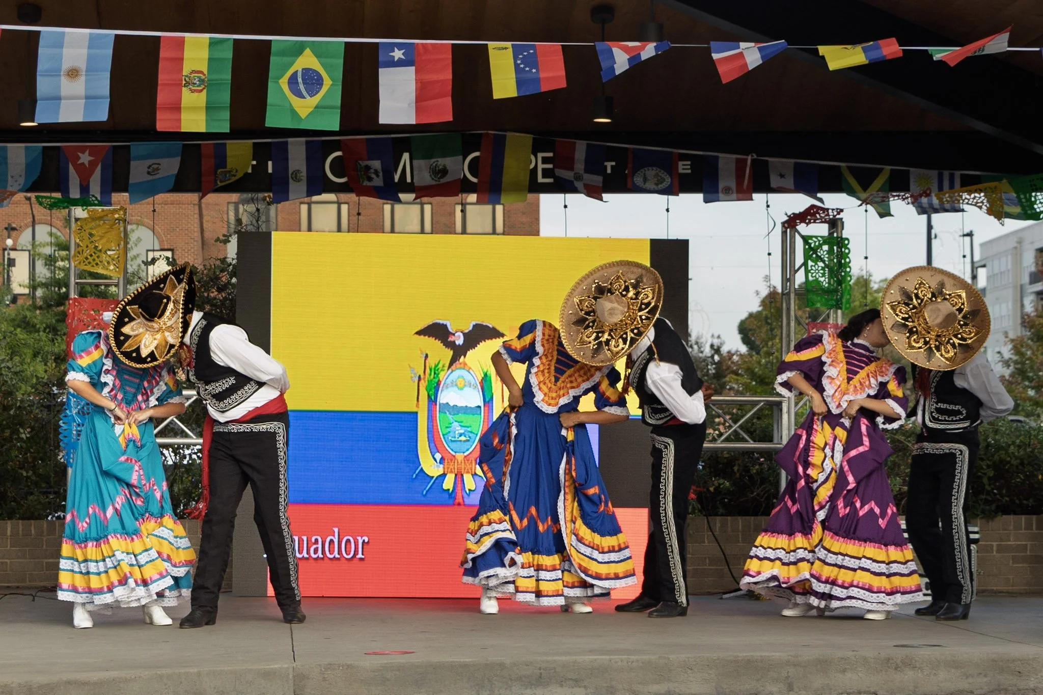 Traditional dancers in colorful costumes performing on stage at a cultural event, with flags of various countries, including Ecuador, hanging above.