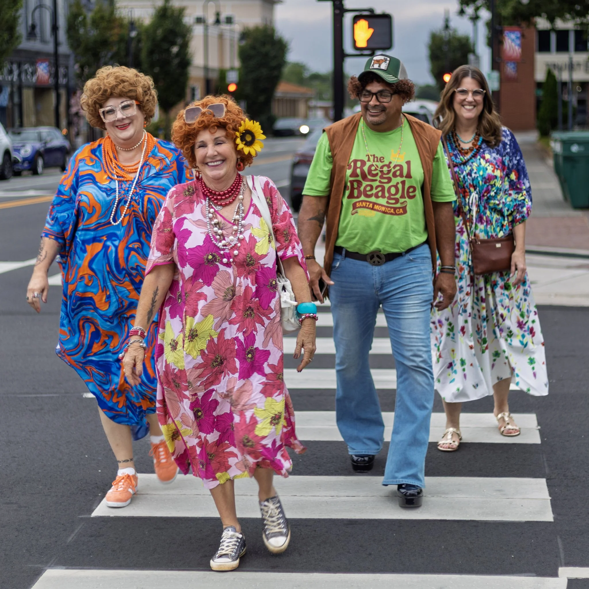 Group of five people dressed in colorful, retro 1960s-style clothing, walking across a crosswalk on a city street. The women are wearing vibrant patterns and accessories, while the man is wearing a green T-shirt with 'The Regal Beagle' text and a pea
