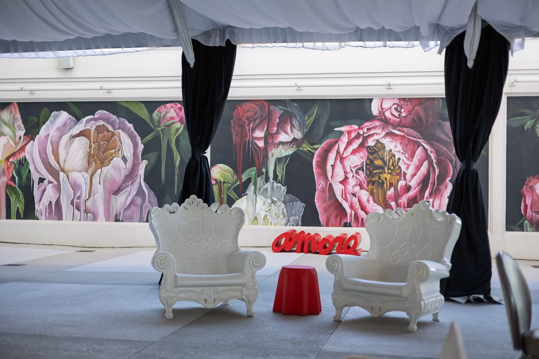 Indoor seating area with two white ornate chairs, a small red table, and a red 'amor' sign in front of a mural of large pink and purple peonies with black curtains hanging from the ceiling.