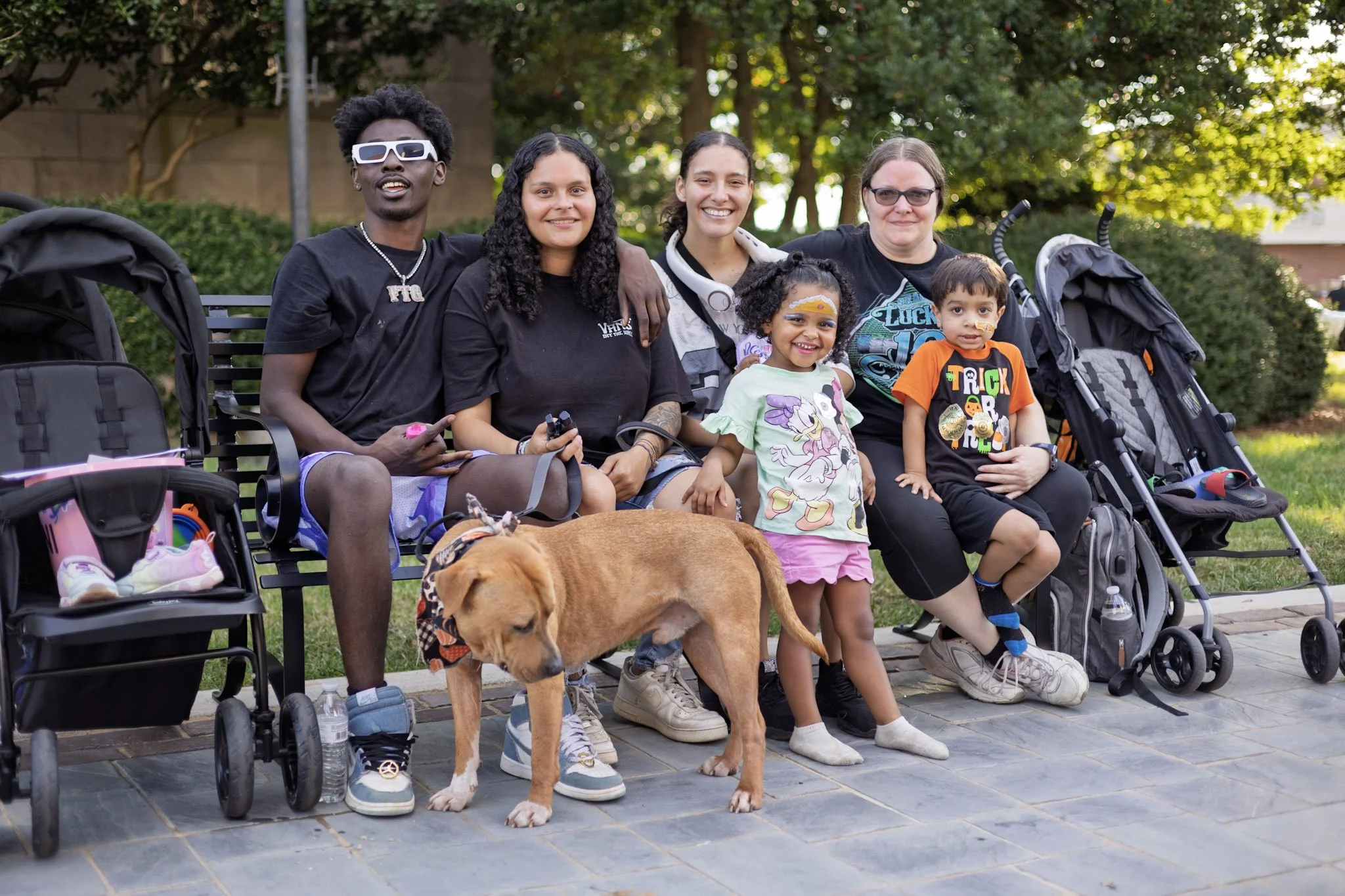 Group of seven people and a dog sitting on a bench in a park during the daytime