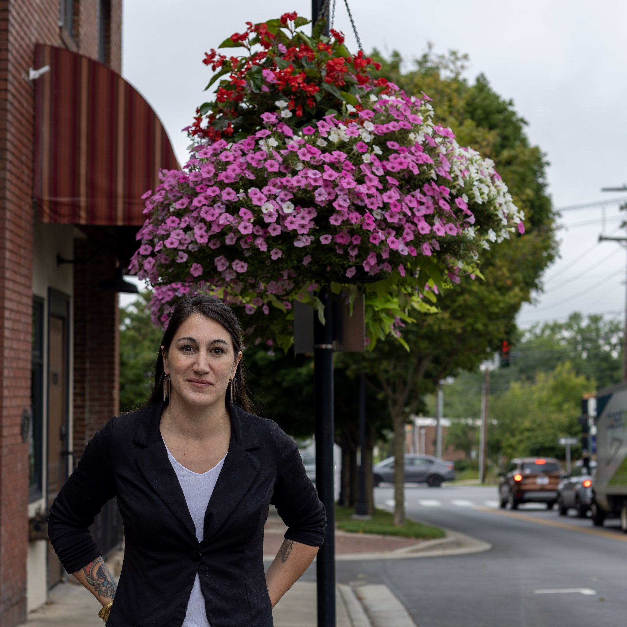 A woman standing on a sidewalk in front of a street with cars and trees, with an overhanging flower basket filled with pink, white, and red flowers above her.