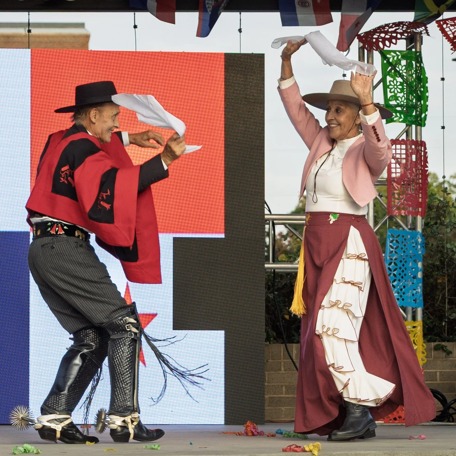 Two women dressed in traditional Mexican attire dancing on a stage with a large screen and colorful papel picado decorations in the background.