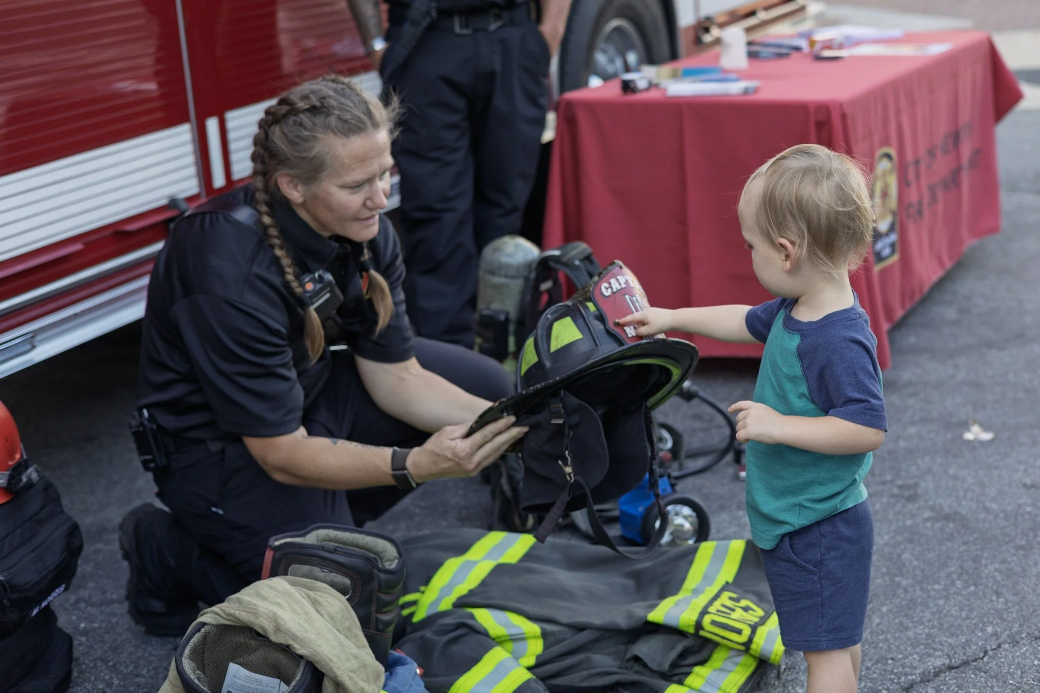 A female firefighter in uniform kneels on the ground, showing her gear to a young boy, who is pointing at her helmet. Firefighting equipment and a red table with informational materials are visible in the background.