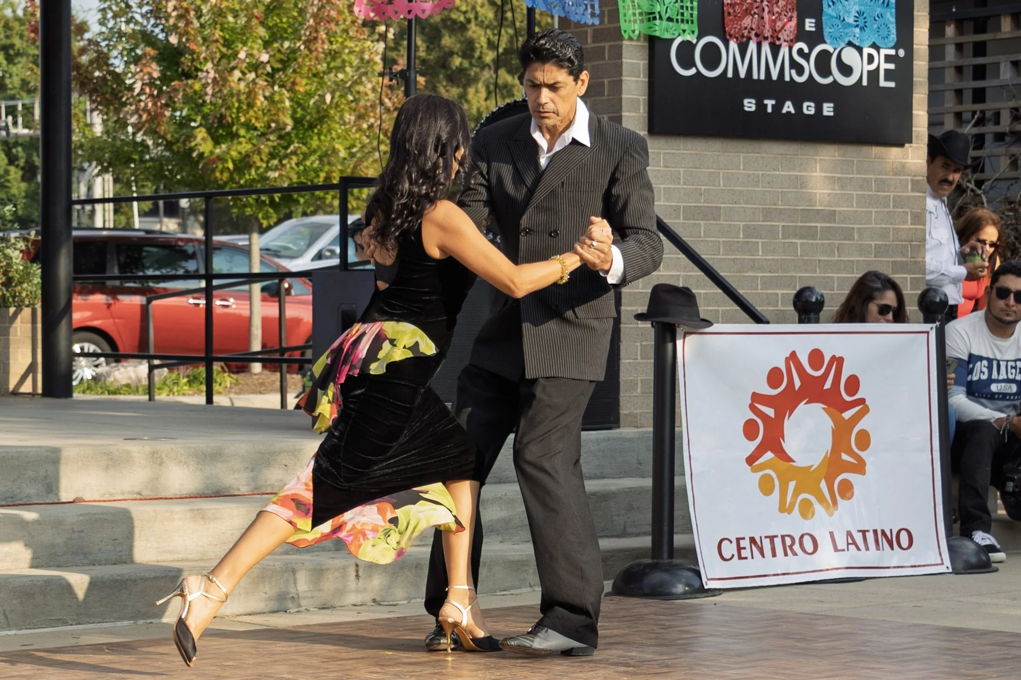 A man and woman dance salsa outdoors on a sidewalk near a comedy club, with onlookers sitting and standing nearby.