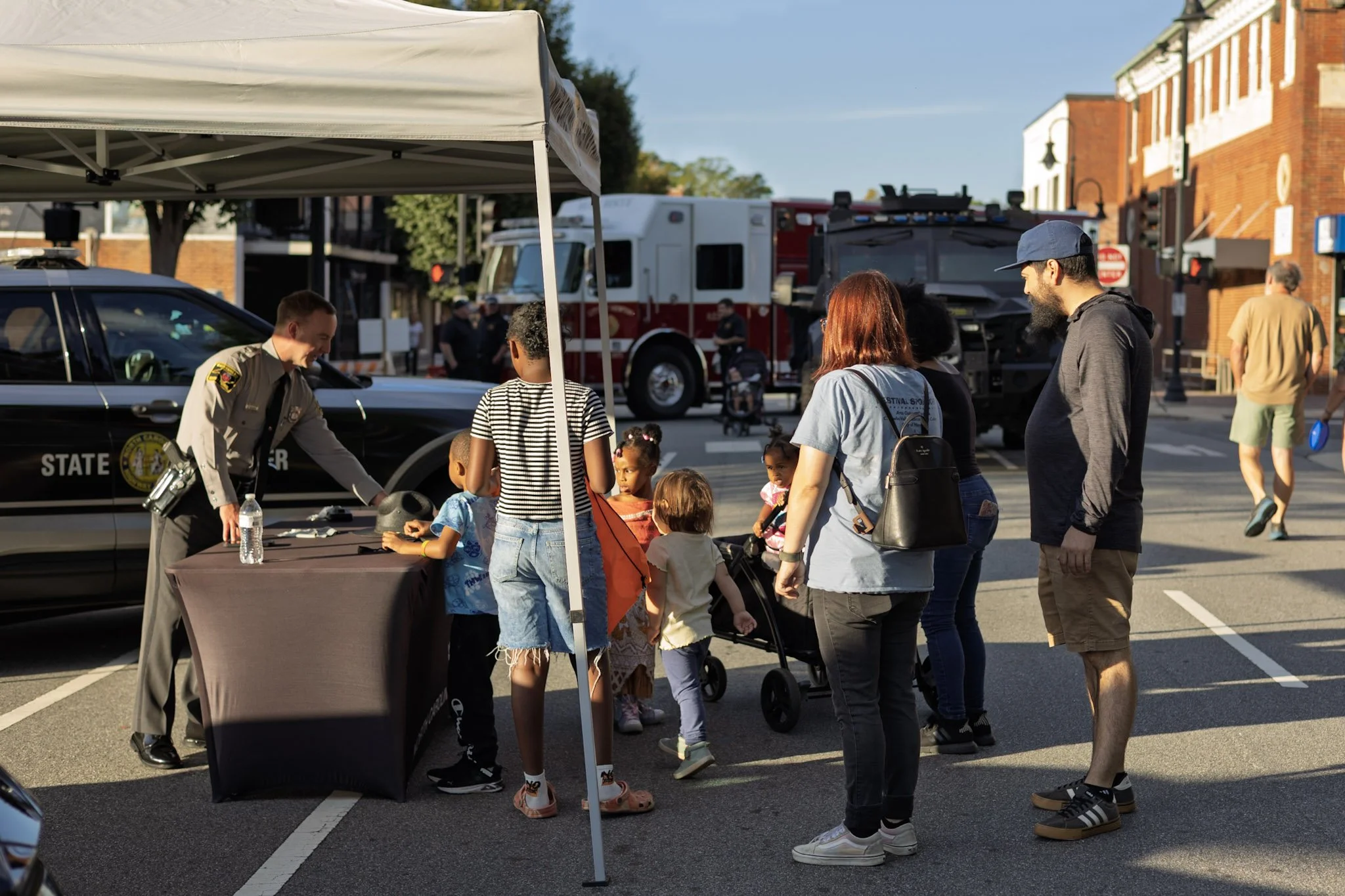 A group of kids and adults gather around a table under a white tent on a city street, interacting with a speaker and water bottle, with police and emergency vehicles in the background.