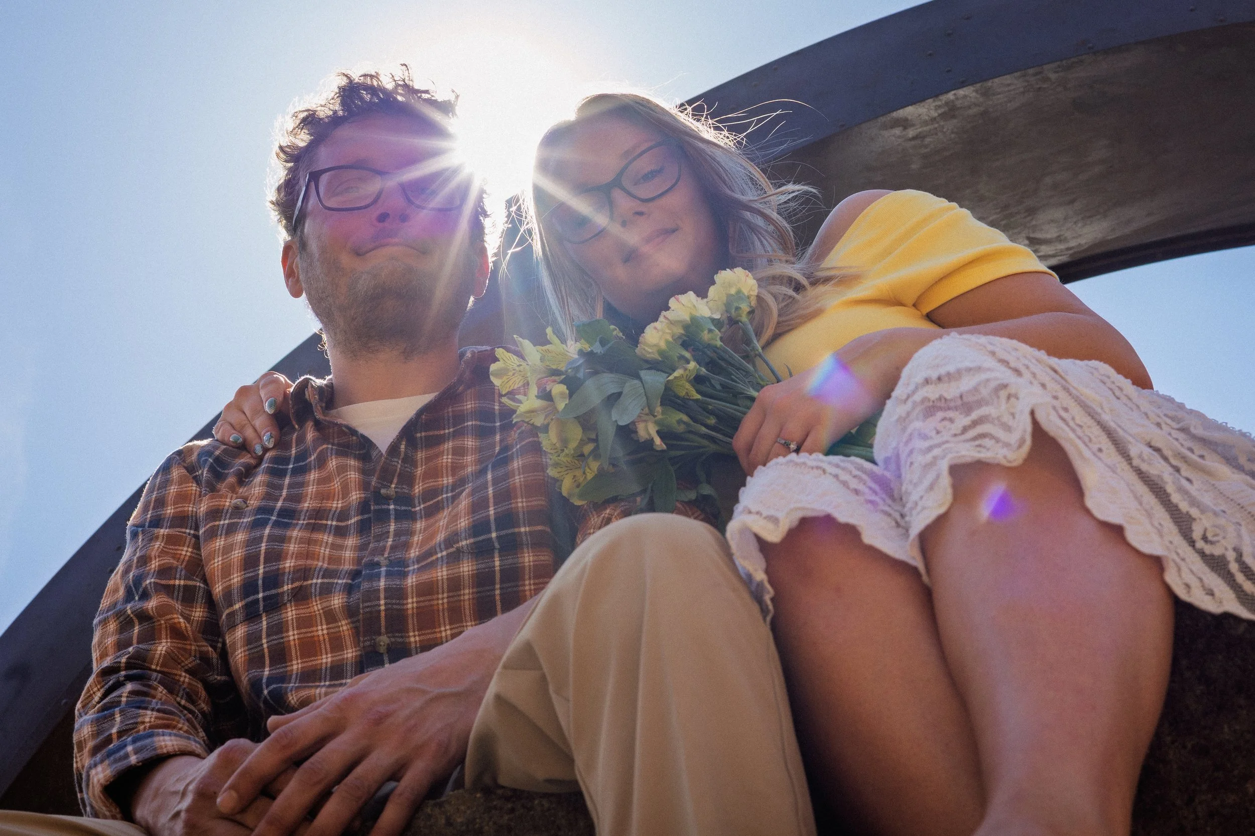 A man and woman are sitting outdoors in bright sunlight, with the sun behind their heads. The man is wearing glasses and a plaid shirt, while the woman is wearing glasses and a yellow top, holding a bouquet of flowers. They are close together, smilin
