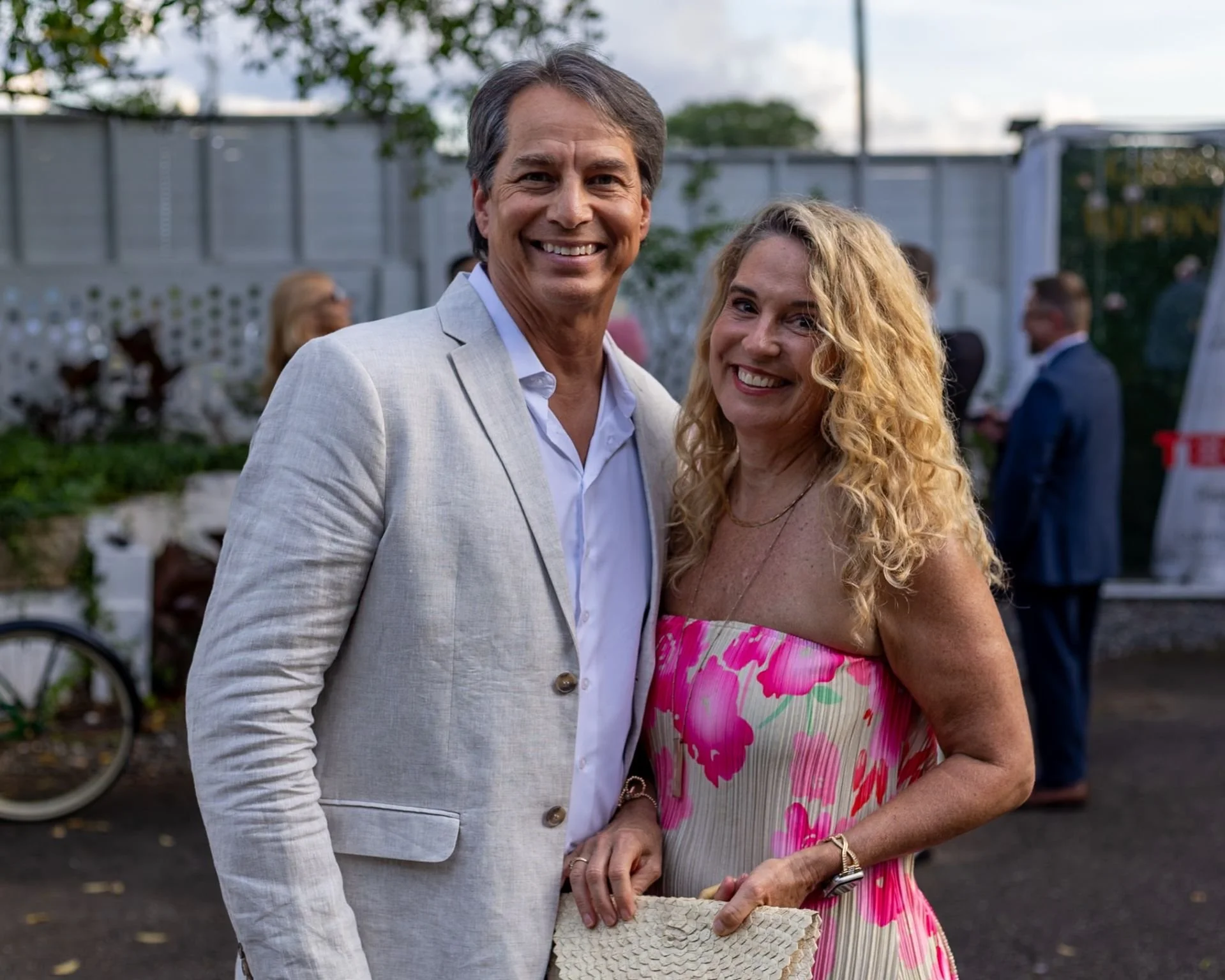 Smiling man and woman at outdoor social event, woman in pink floral dress holding a woven clutch purse, man in light blazer, background with other people and a tent.