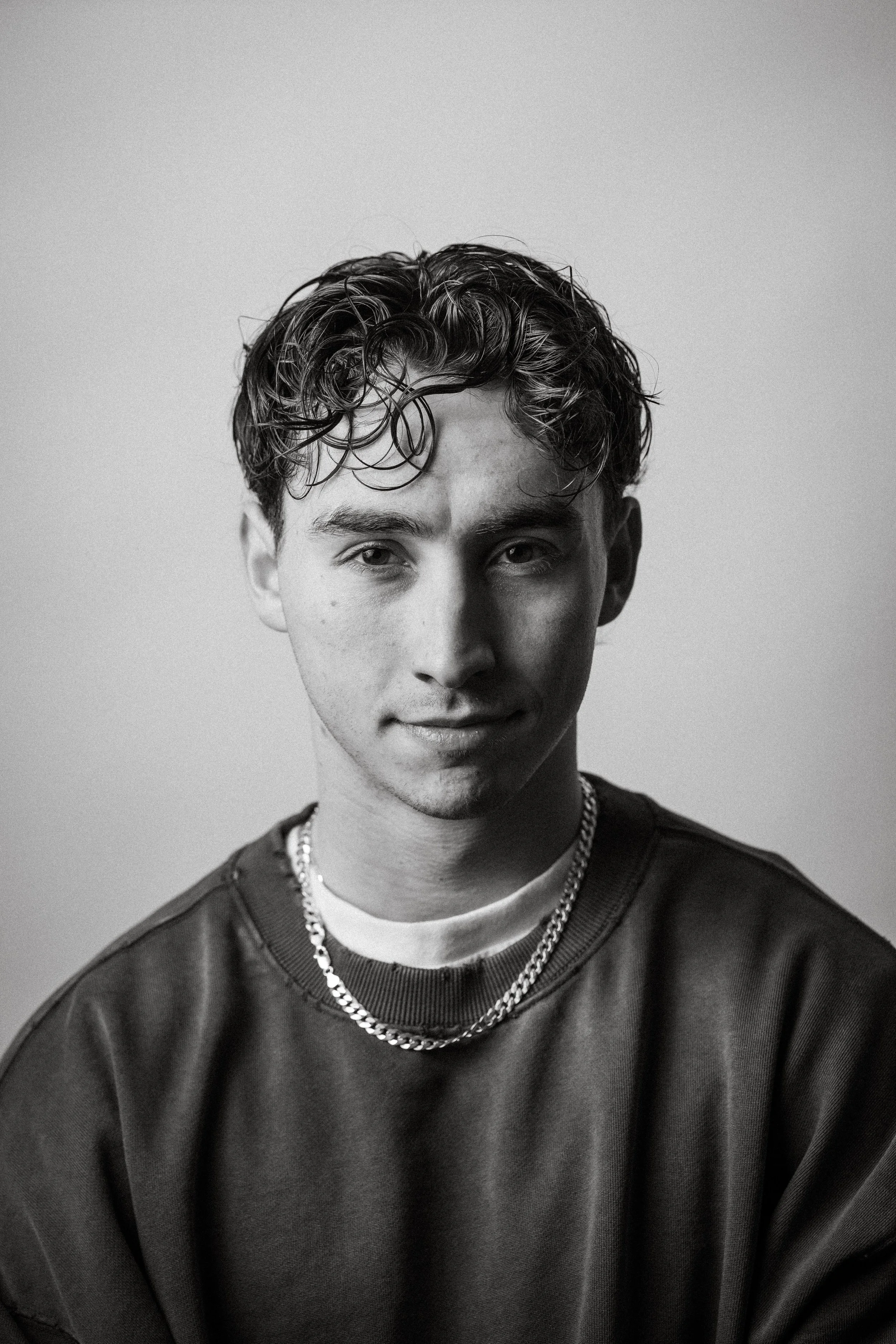 Black and white portrait of a young man with wavy hair, wearing a dark sweatshirt, a chain necklace, and a white undershirt, against a plain background.