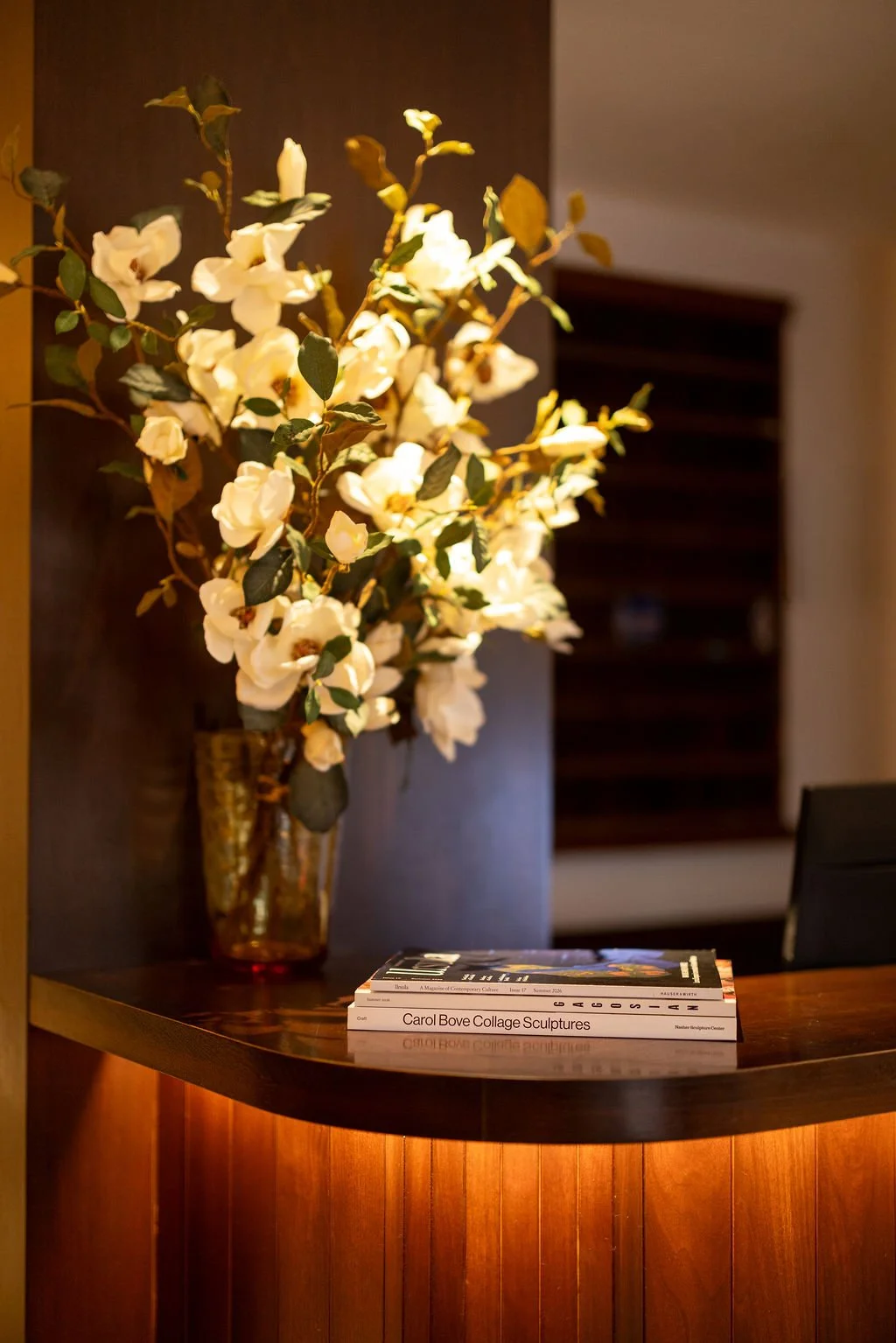 Decorative white flower arrangement in a gold-colored vase on a wooden table, with magazines underneath, against a dark background.