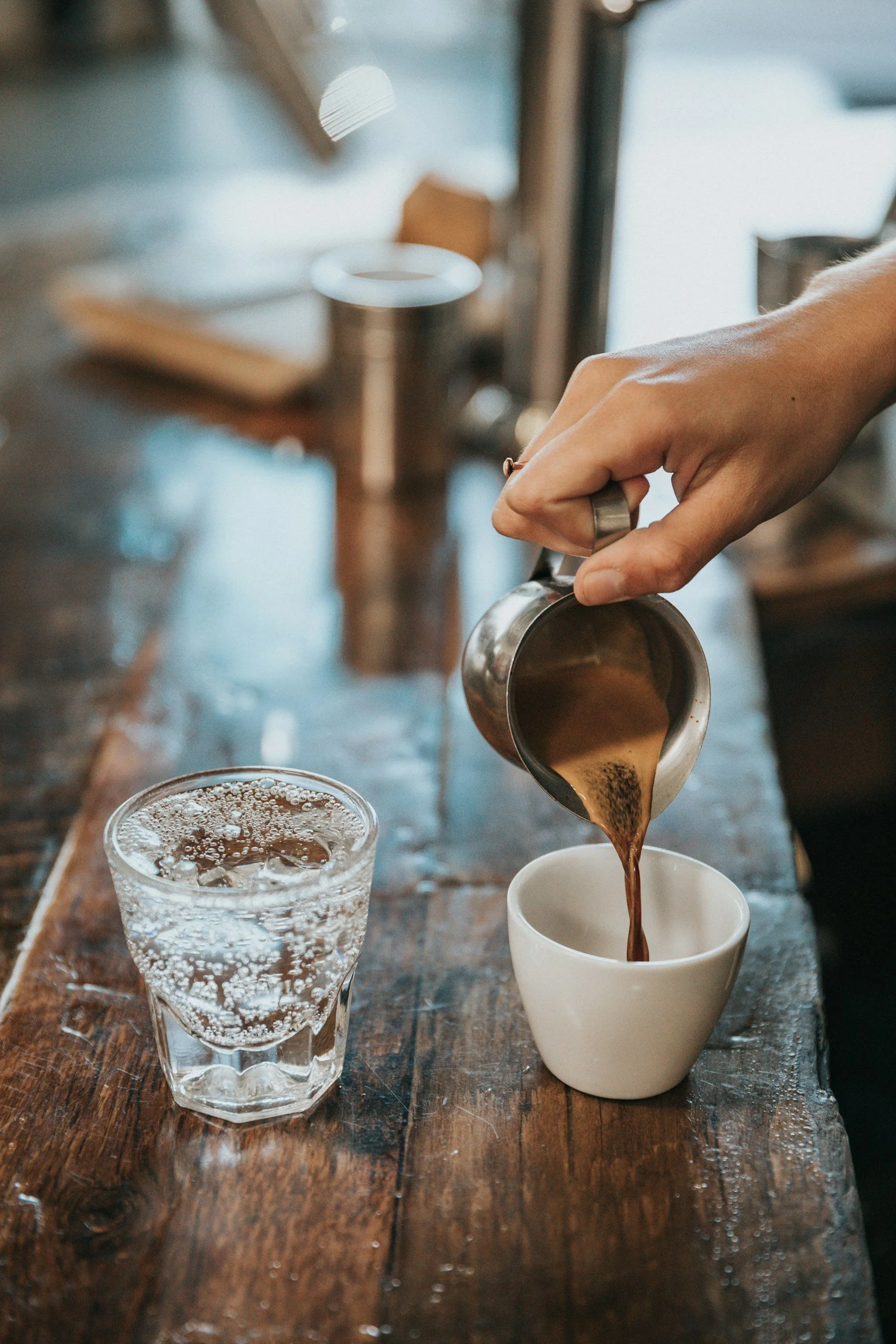 A person pouring coffee from a metal pitcher into a white ceramic cup on a wooden table. A glass of water is beside the cup, and the background is blurred with a cozy indoor setting.