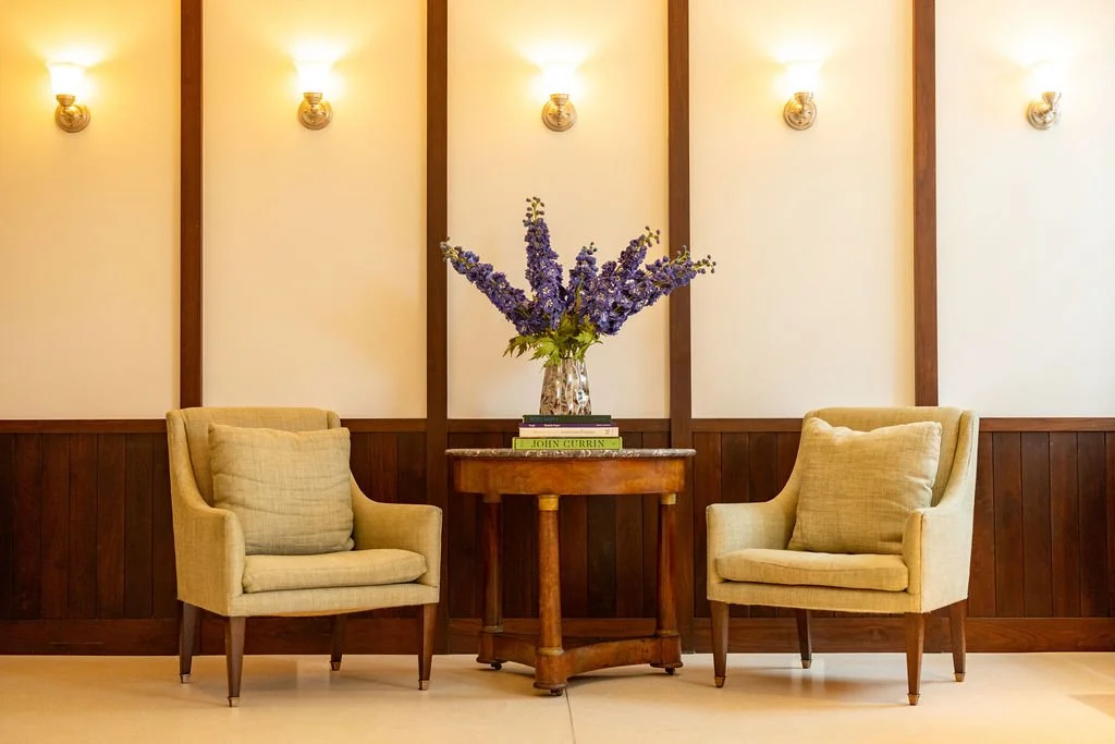 A cozy sitting area with two beige armchairs flanking a small wooden table with a glass vase of purple flowers and a few books inside. The wall behind has warm lighting fixtures and wood paneling.
