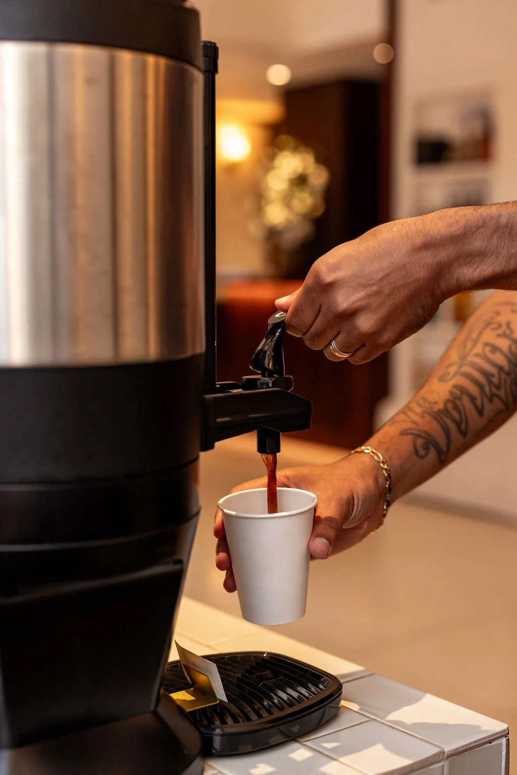A person with tattoos on their arm is filling a white paper cup with coffee from a large, stainless steel coffee dispenser.