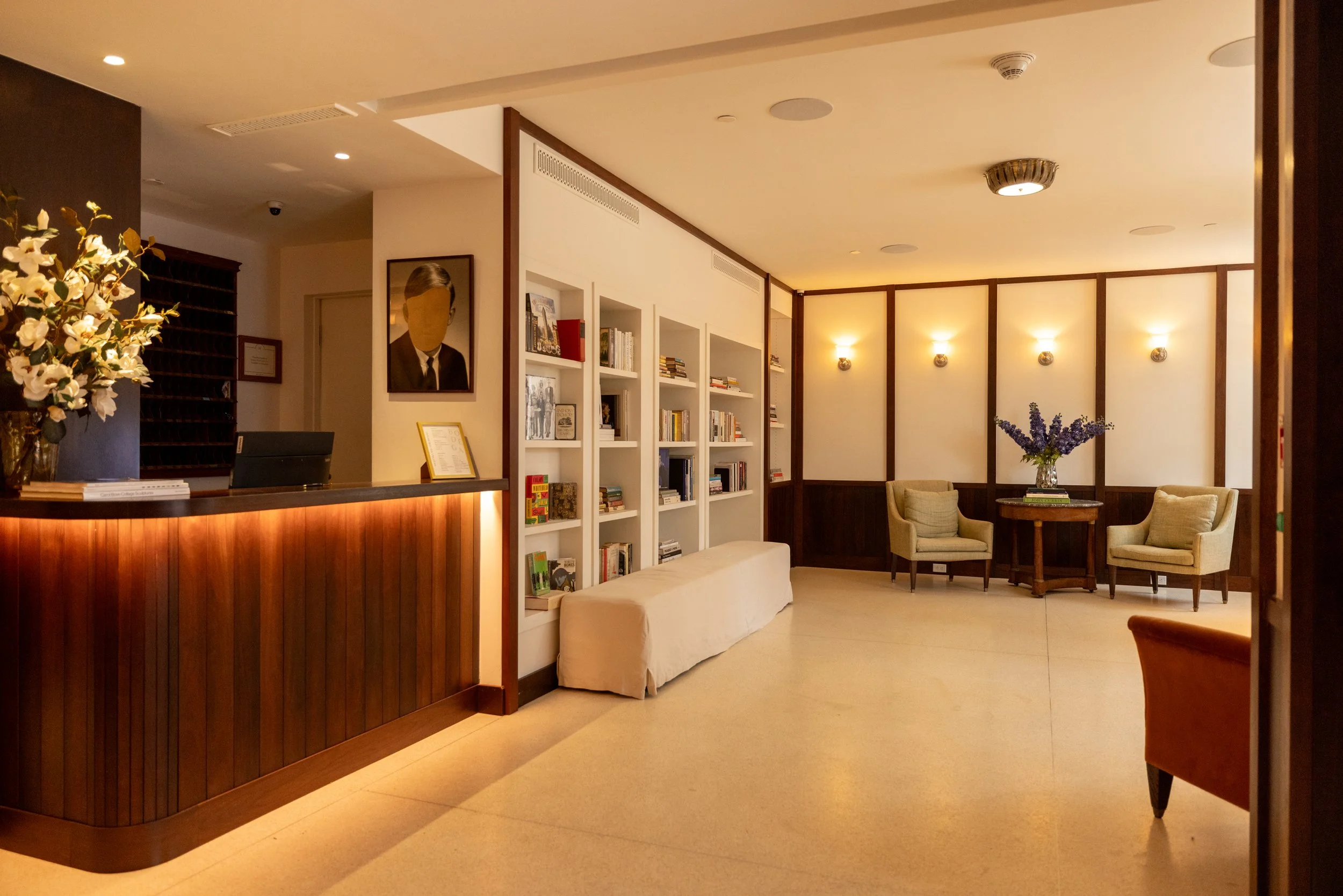 A cozy hotel lobby with seating area, bookshelf, framed art, and floral decor.