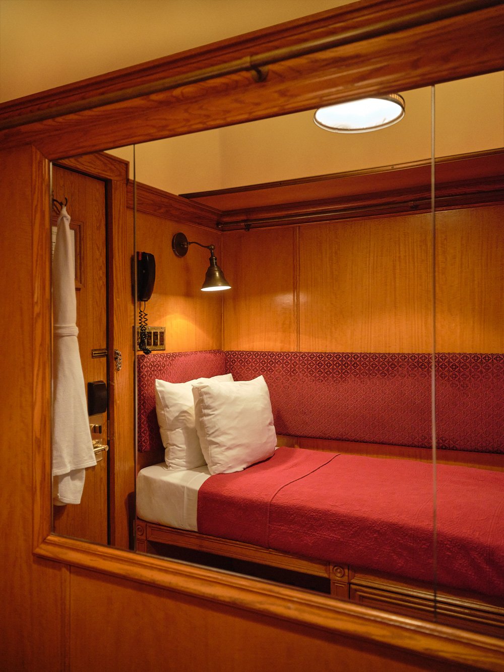 A cozy hotel room with wood-paneled walls, a single bed with white pillows and red bedspread, and a mirror reflecting the room’s interior.