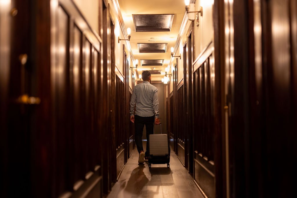A man walking in a narrow, dimly lit hallway with wooden paneling, pulling a suitcase behind him.