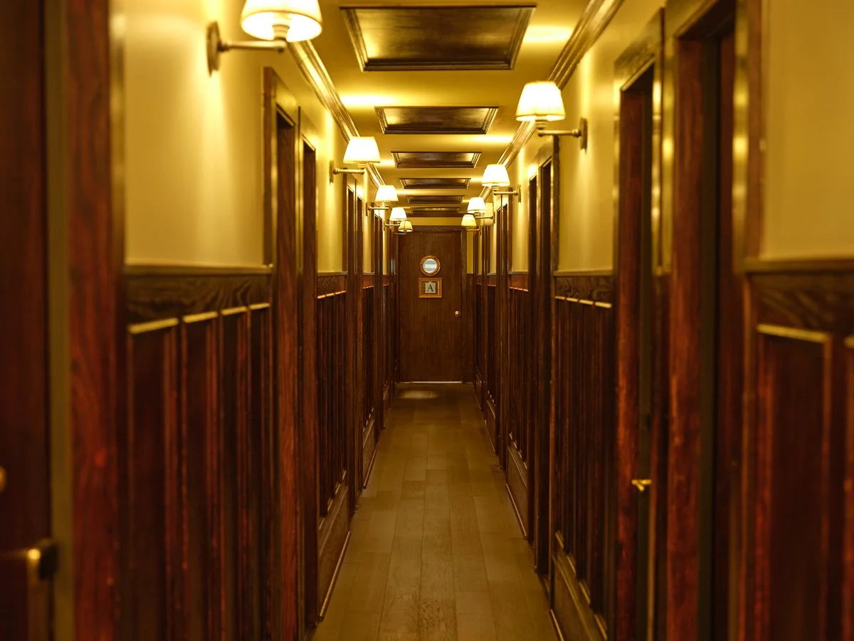 A narrow hotel corridor with wooden paneling and multiple wall-mounted lamps along the ceiling.