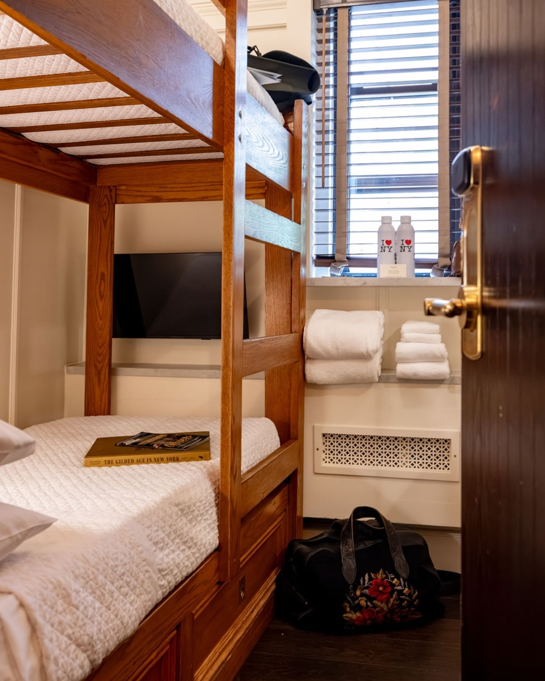 A small room with a wooden bunk bed, a flat-screen TV, folded white towels on a shelf, two bottles with 'I ♥ NY' on the windowsill, and a black bag with floral embroidery on the floor.