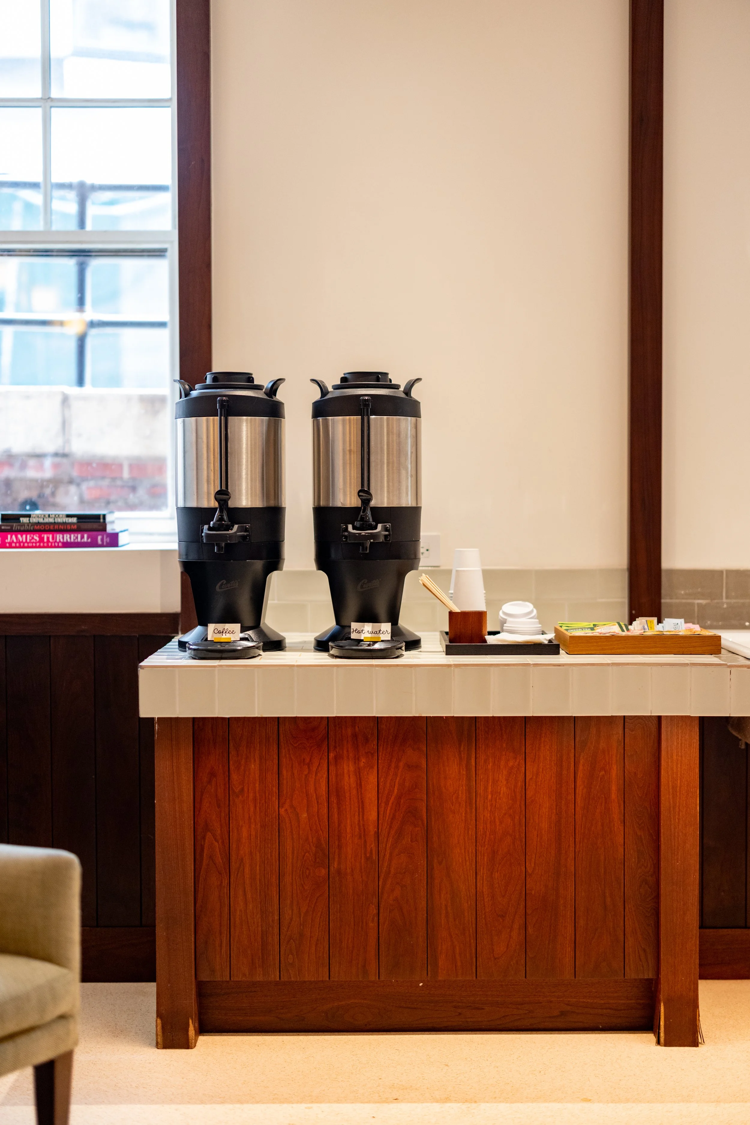 Coffee station with two large coffee dispensers, disposable cups, stir sticks, and snacks on a wooden countertop in a cozy room with a window and beige carpet.