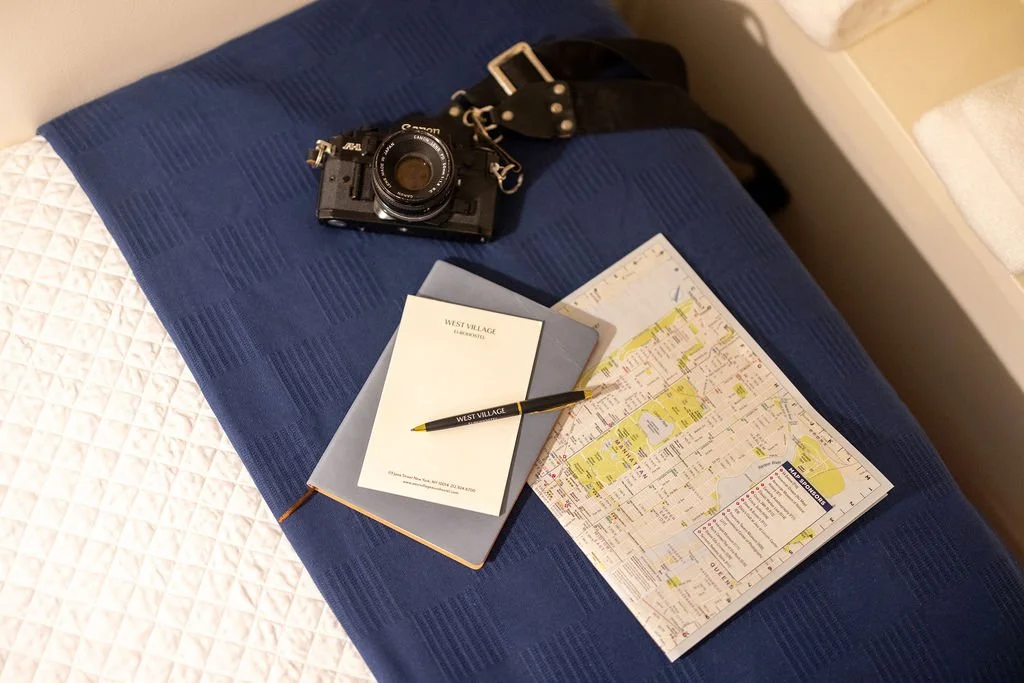 A camera, a travel guide, a notepad with a pen, and a map laid out on a blue bedspread in a hotel room.