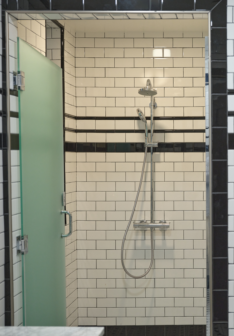 A shower with white subway tiles, black border tiles, a stainless steel shower head, and a separate handheld shower attachment. Part of a frosted glass door is visible on the left.