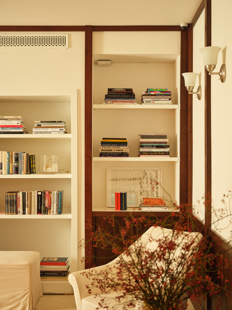 Interior of a room with white bookshelves filled with books, a white armchair with a floral arrangement in front, and framed artwork on one shelf.