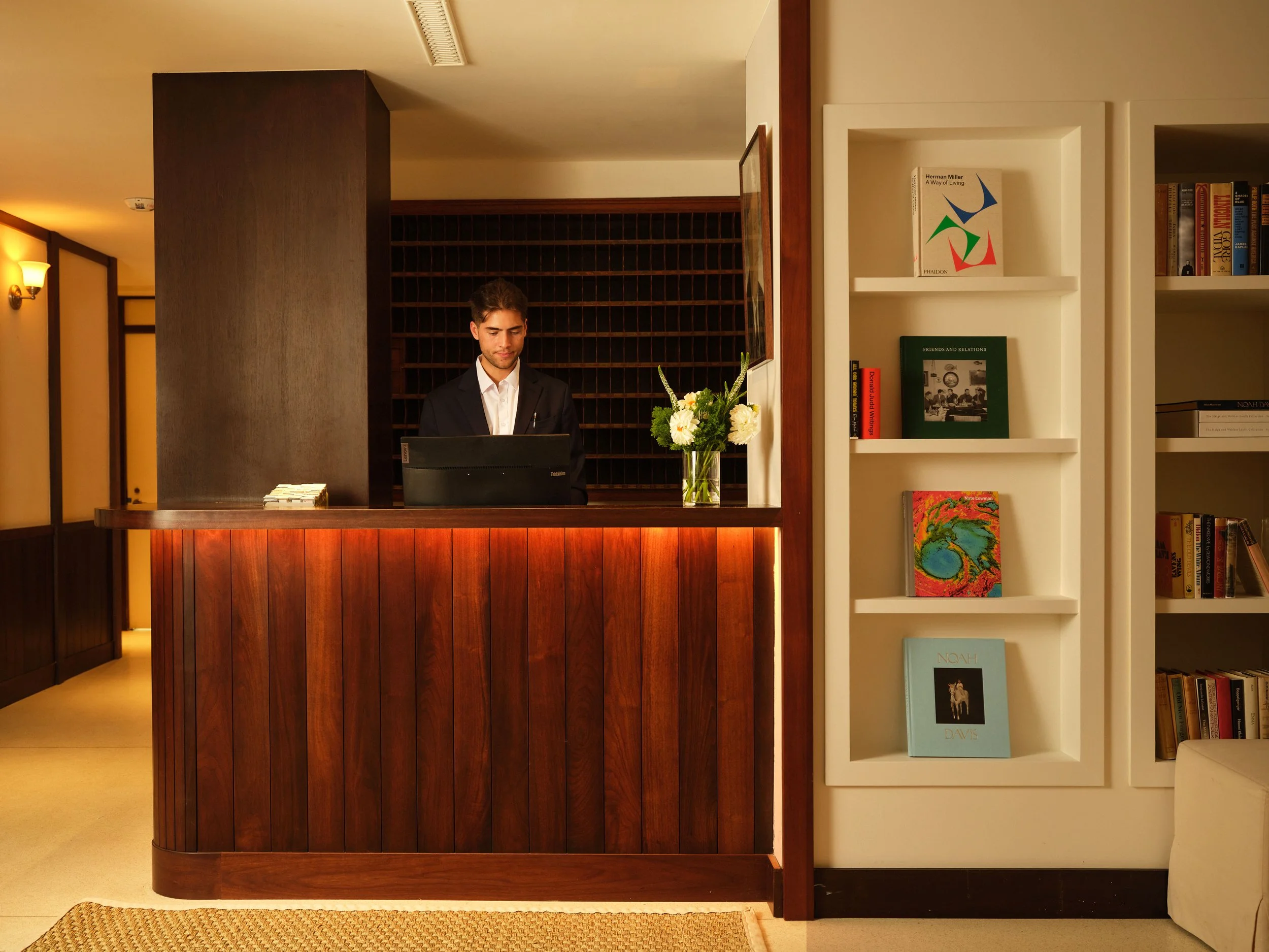 A hotel reception desk with a young man in a suit standing behind it, decorated with a bouquet of white flowers, gold wall sconces, and warm lighting. To the right, a white bookshelf holds books and artwork.