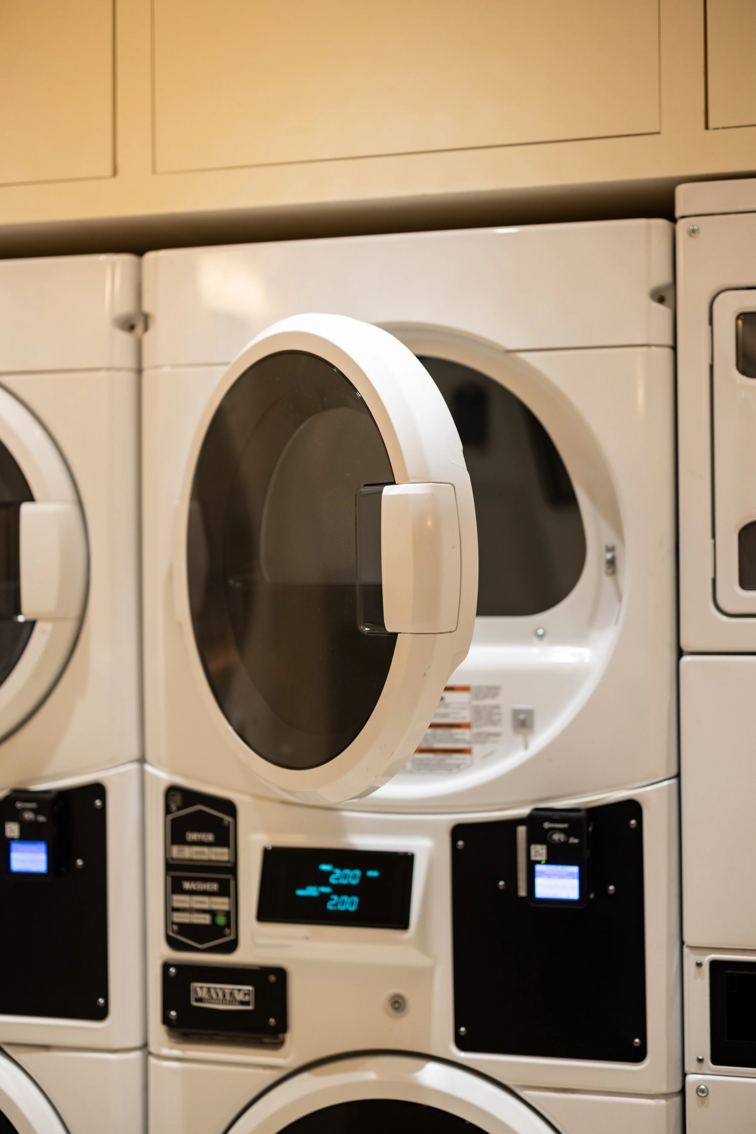 A stack of laundry washing machines and dryers, with one dryer door slightly open showing the interior, in a laundromat.