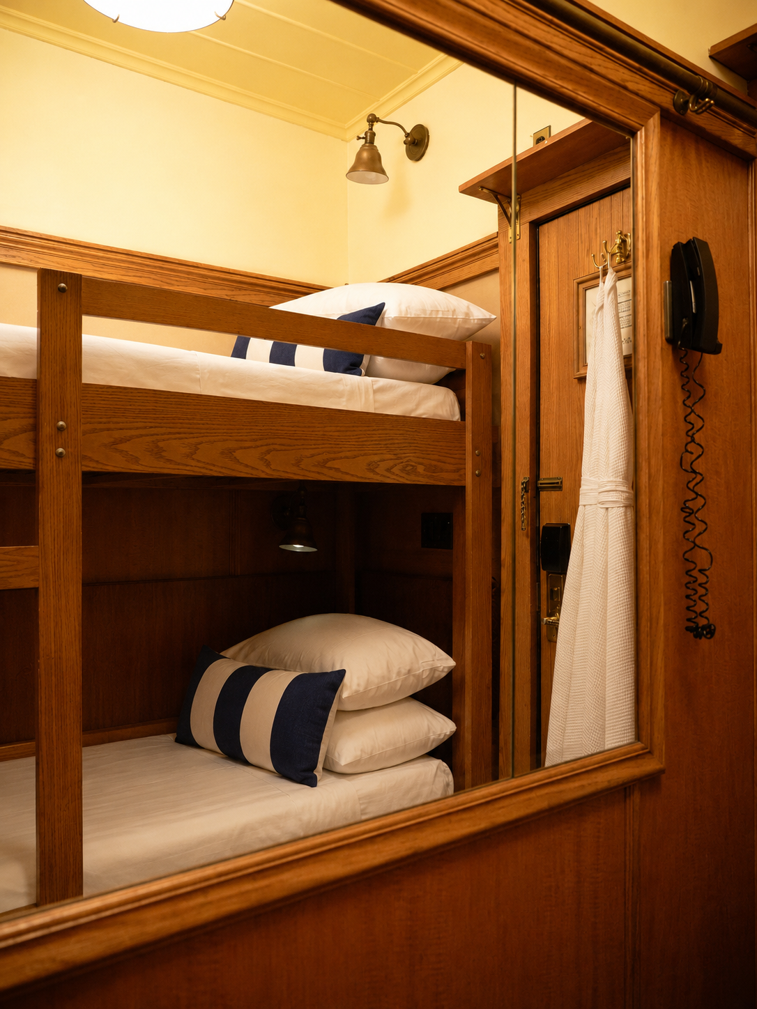 A set of wooden bunk beds with white bedding and navy blue and white striped pillows, inside a warm wood-paneled room with a mirror, a wall-mounted telephone, a robe hanging on a hook, and soft warm lighting.