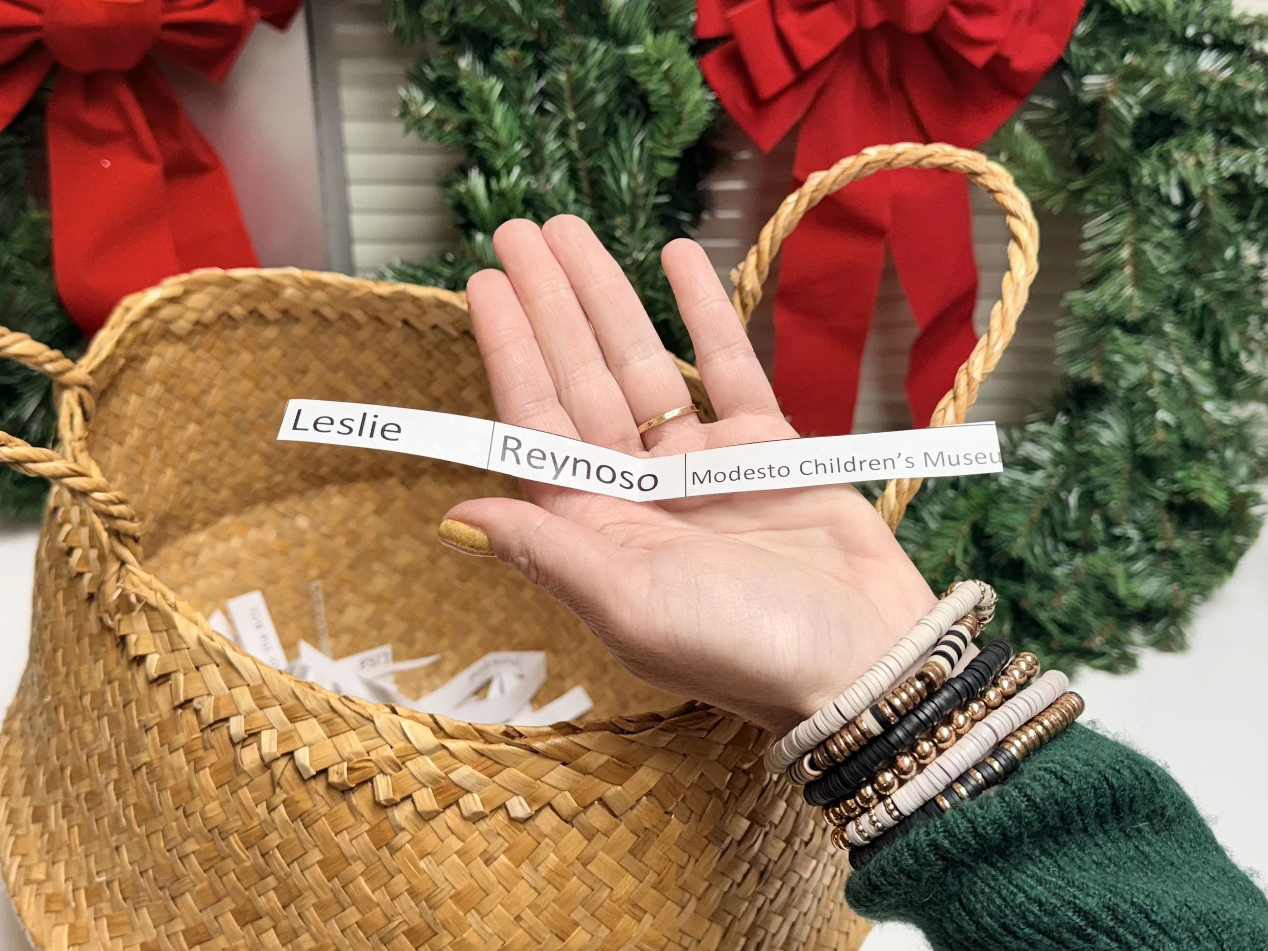 A wicker basket filled with Christmas presents wrapped in brown paper with green ribbons, decorated with pinecones, red baubles, a candy cane, a yellow gift bow, a star-shaped decoration, and green pine branches.