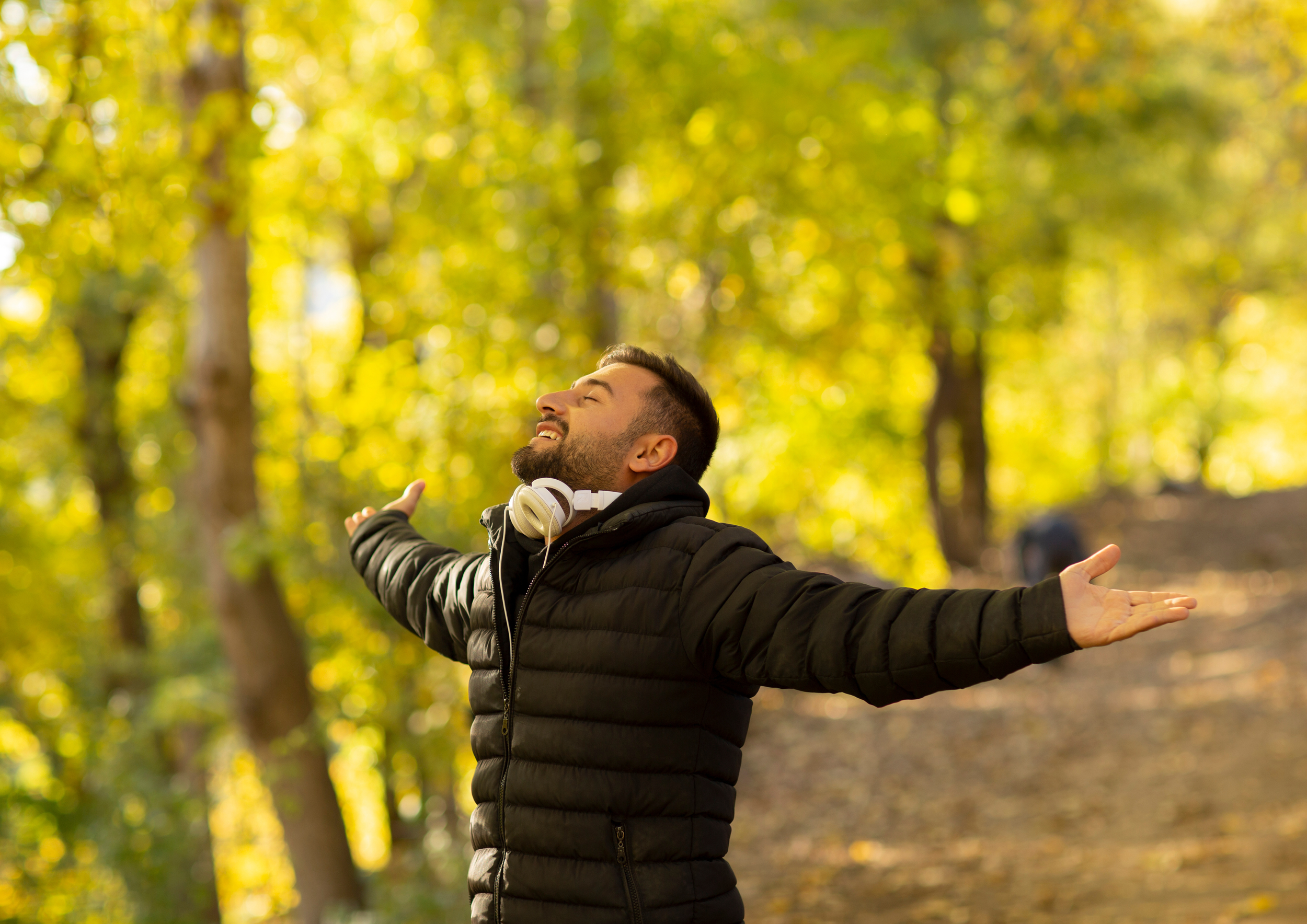 A joyful man with headphones around his neck, wearing a black puffer jacket, standing with arms outstretched in a sunlit forest during autumn.
