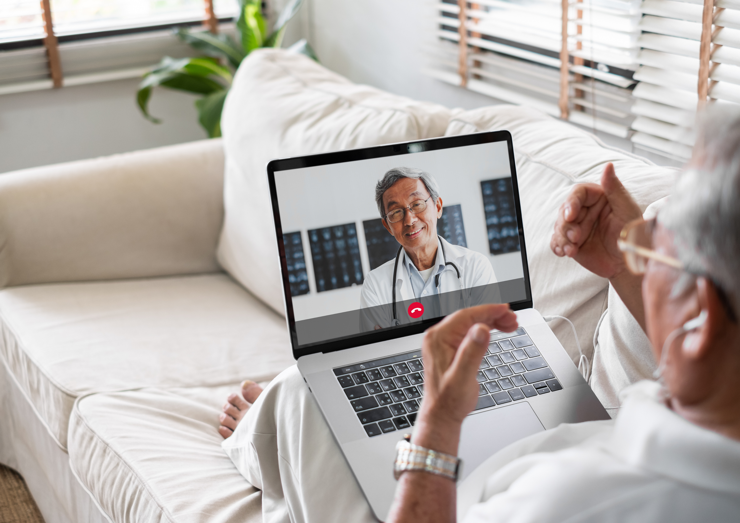 An elderly woman having a video call with a male doctor on a laptop, sitting on a white sofa in a bright living room.