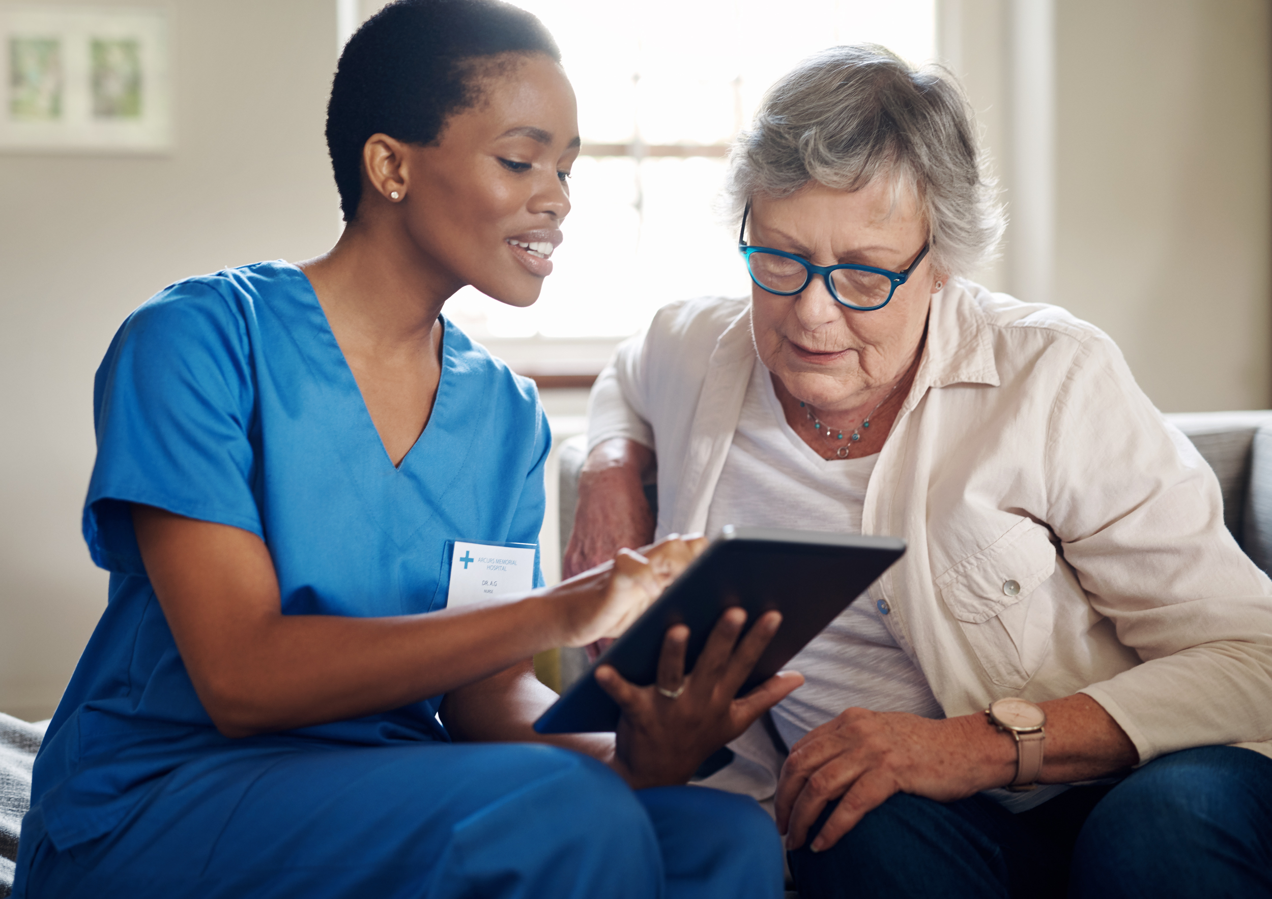 A nurse in blue scrubs explaining a digital tablet to an elderly woman sitting on a couch.