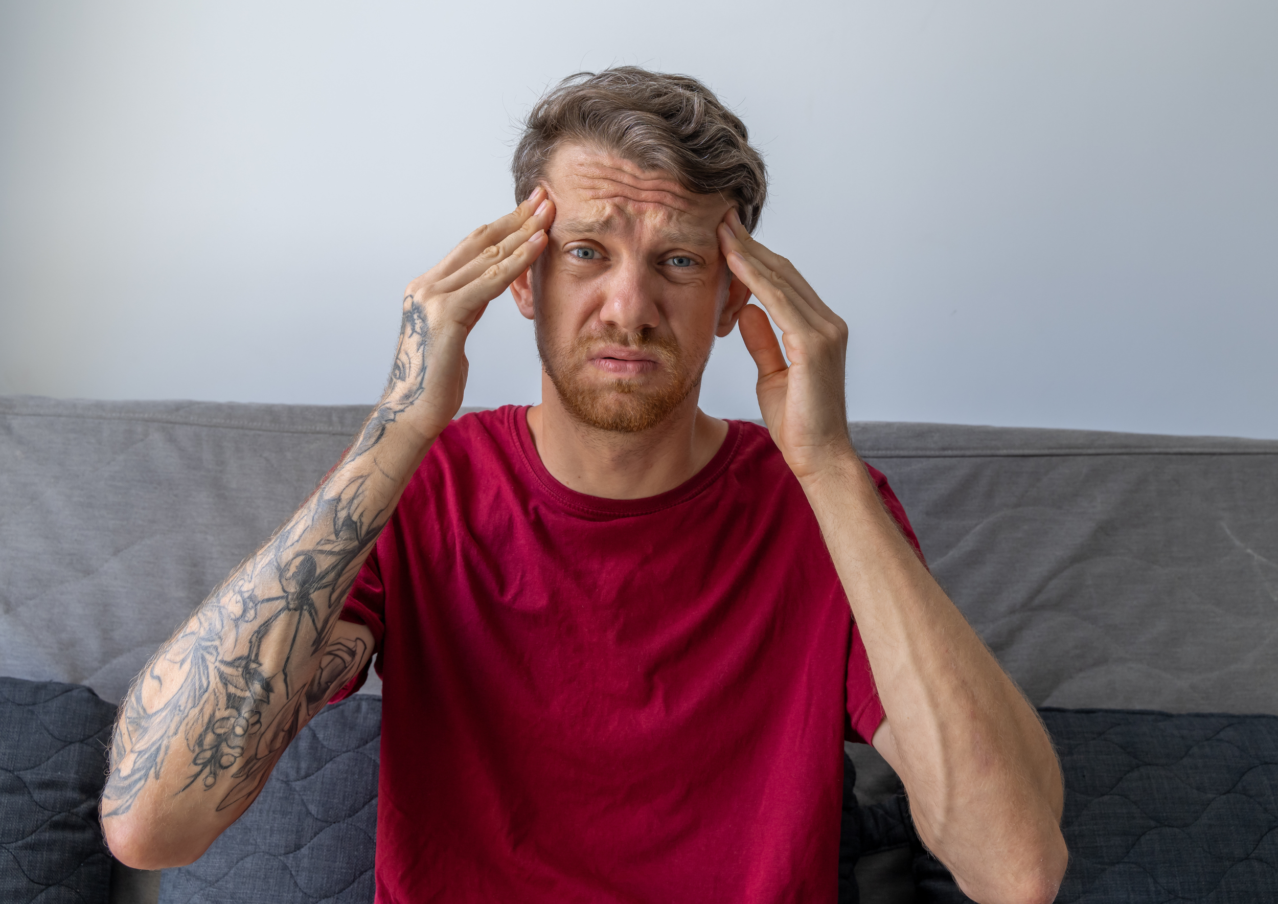 A man with short brown hair, light skin, and tattoos on his left arm, wearing a red T-shirt, sitting on a grey couch. He is holding his forehead with both hands, looking distressed or confused.