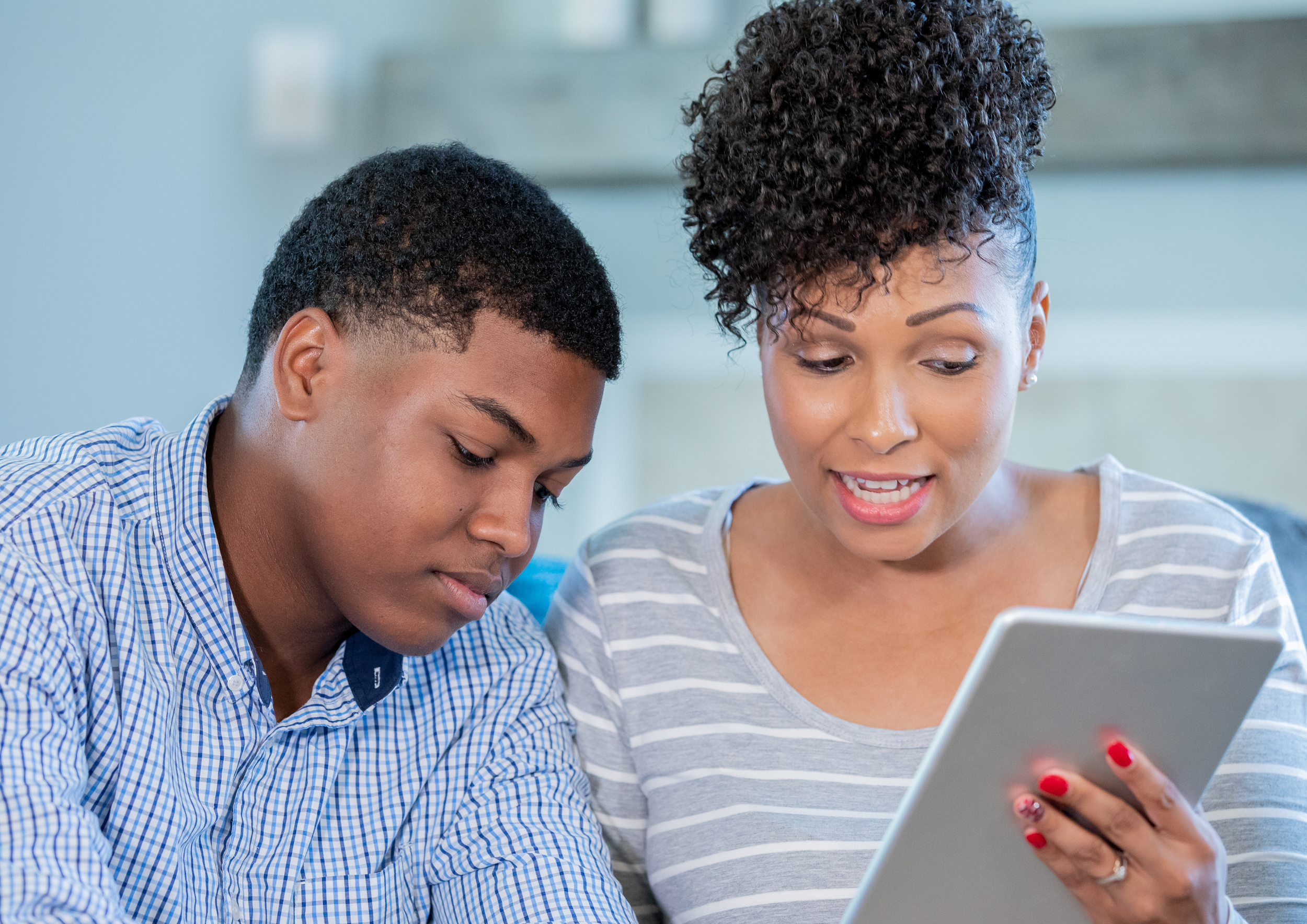 A woman and a boy sitting close together indoors, looking at a tablet device. The woman is explaining something to the boy, who is attentively listening.