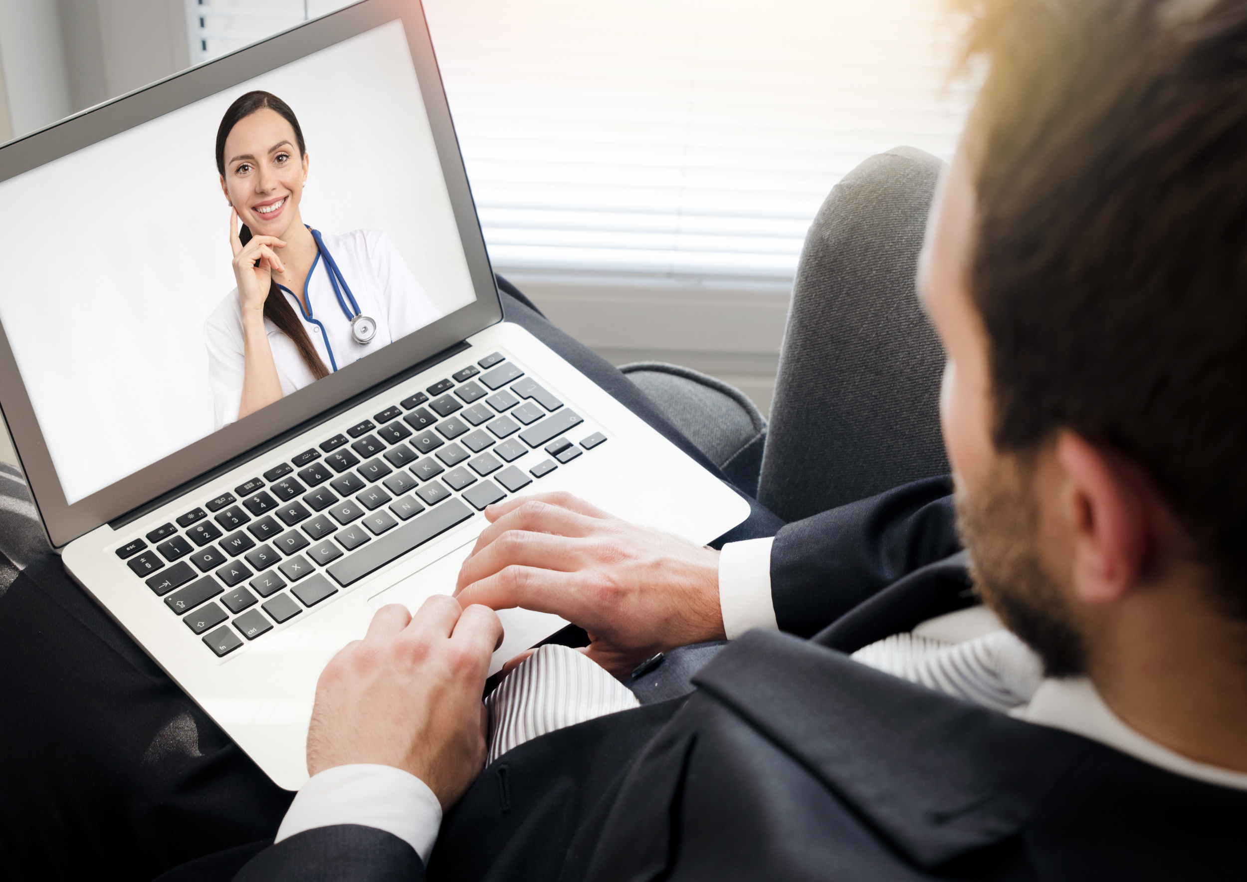 A man in a suit on a video call with a female doctor on a laptop screen in a bright room.