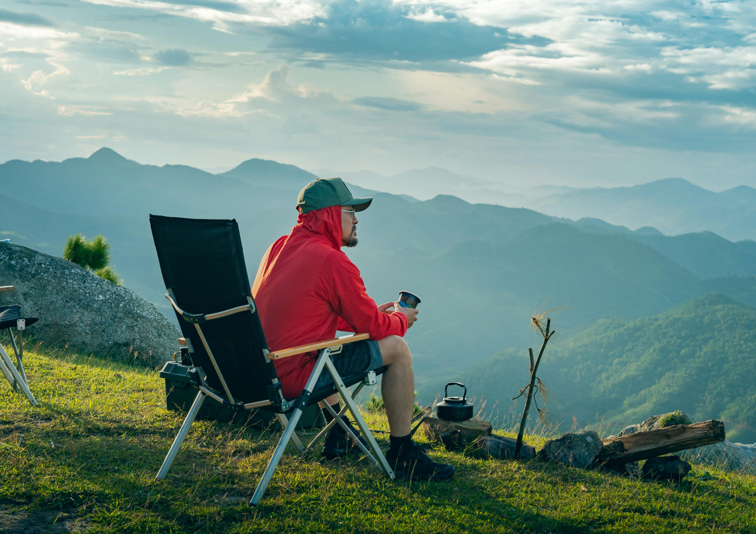 A man sitting on a camping chair on a grassy hilltop, holding a cup, overlooking a mountain range during daylight.