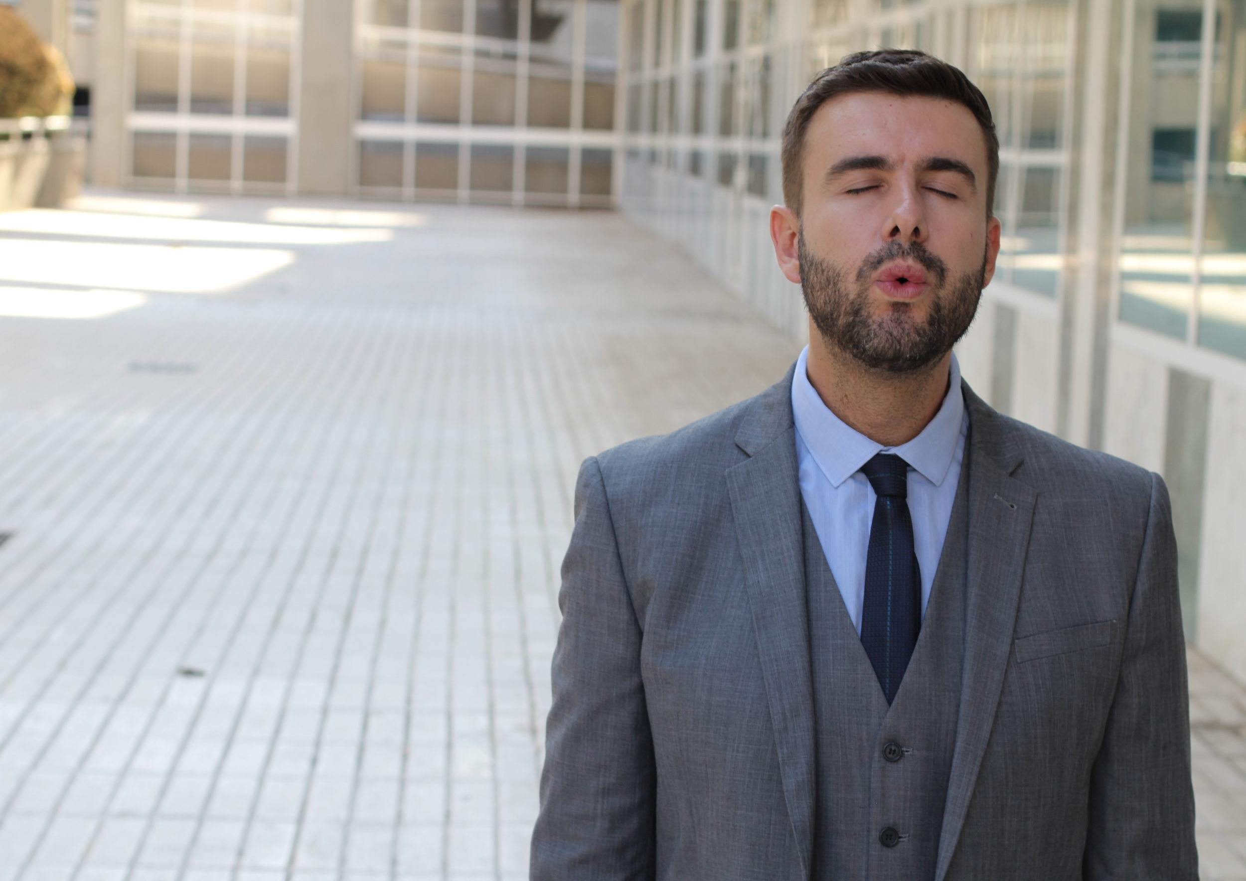 A man in a gray business suit with a blue shirt and dark tie stands outdoors with his eyes closed, lips pursed as if blowing a kiss or whistling, on a paved area in front of a modern building with glass windows.