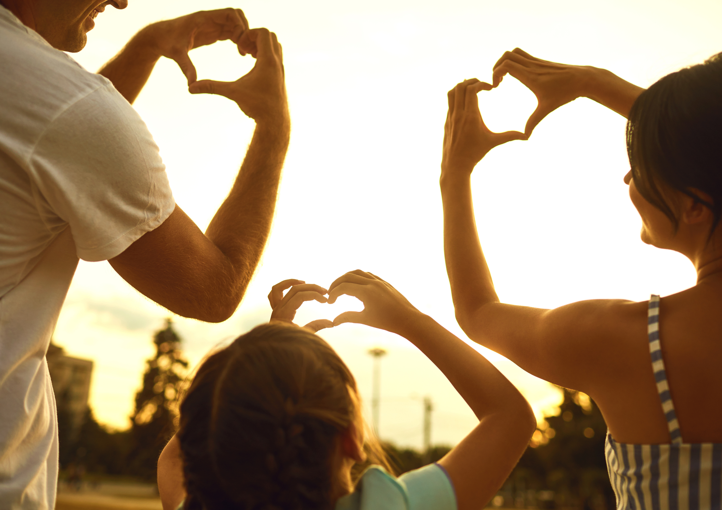 A family making heart shapes with their hands during sunset, with trees and buildings in the background.