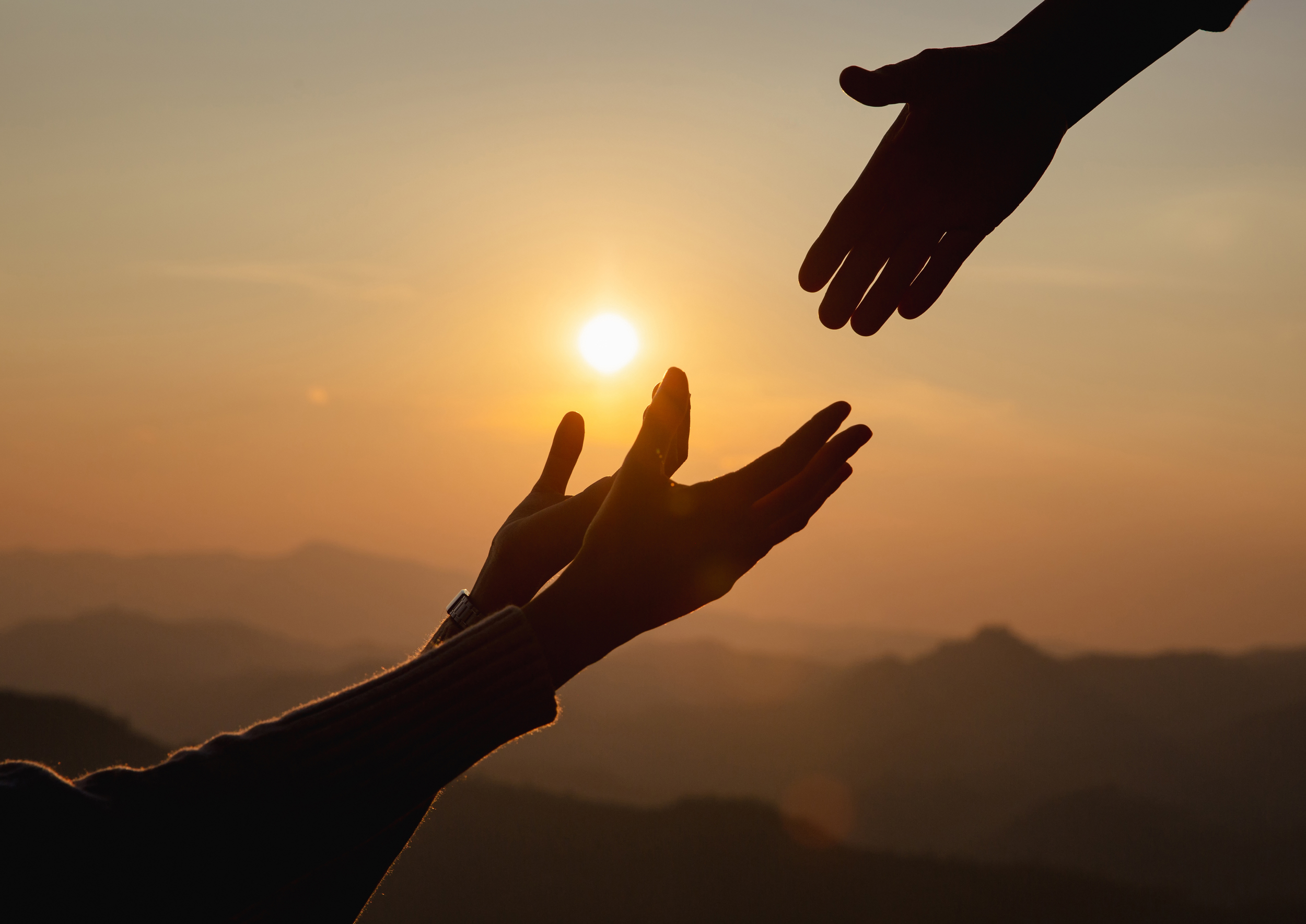 Silhouettes of two hands reaching towards each other with the setting sun between them, against a mountainous horizon at sunset.