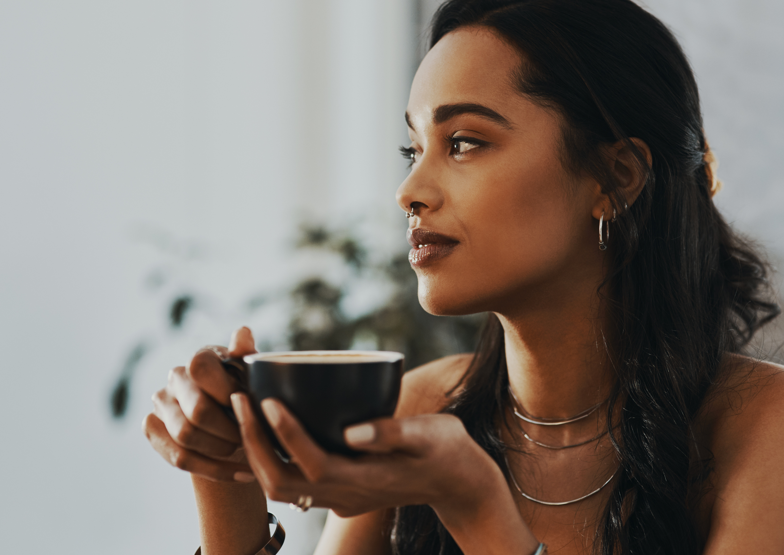 A woman with dark hair and jewelry holding a black coffee cup and looking to the right, indoors with a blurred background.