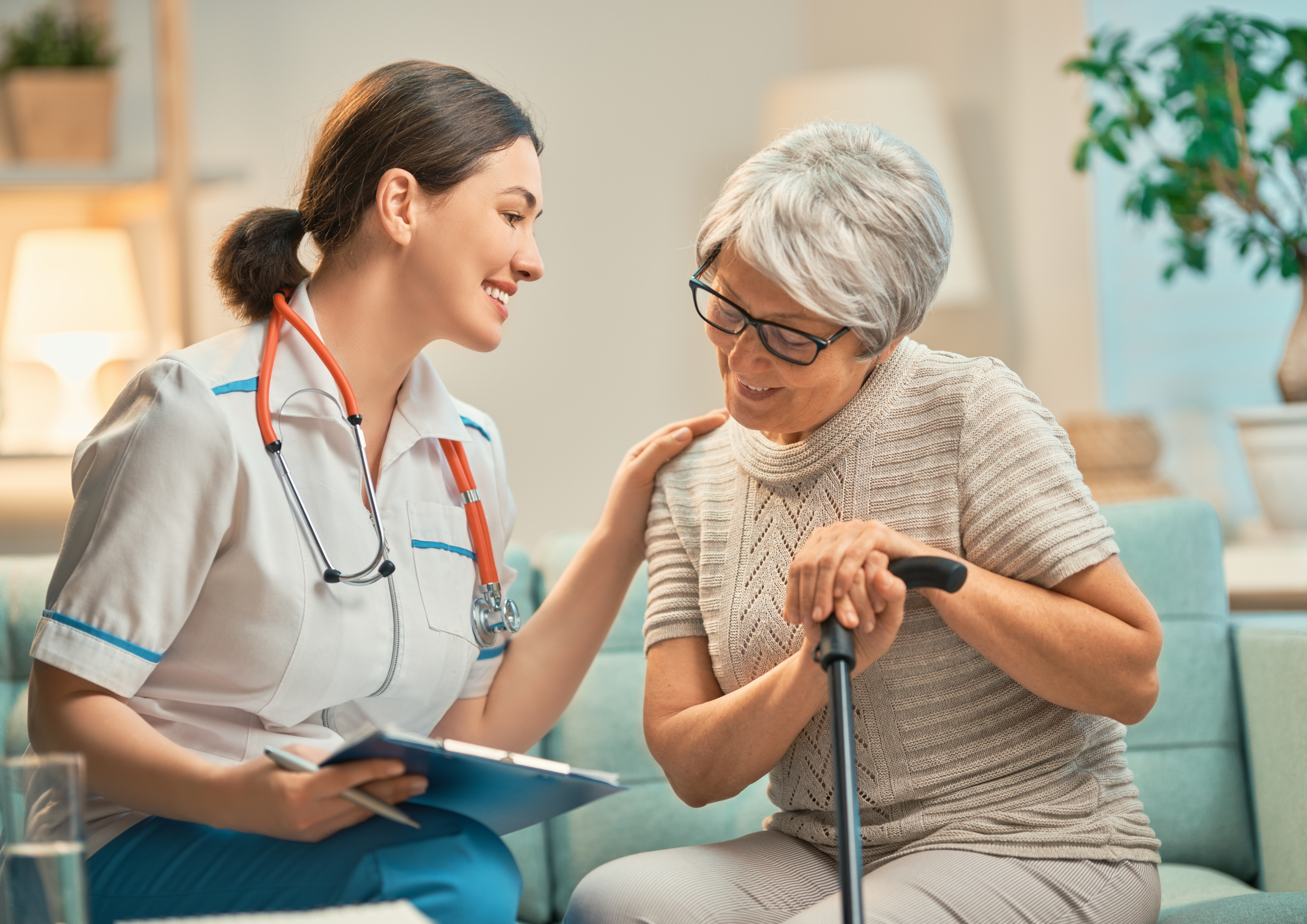 A young female nurse smiling and talking to an elderly woman with a walking cane in a cozy, well-lit living room.