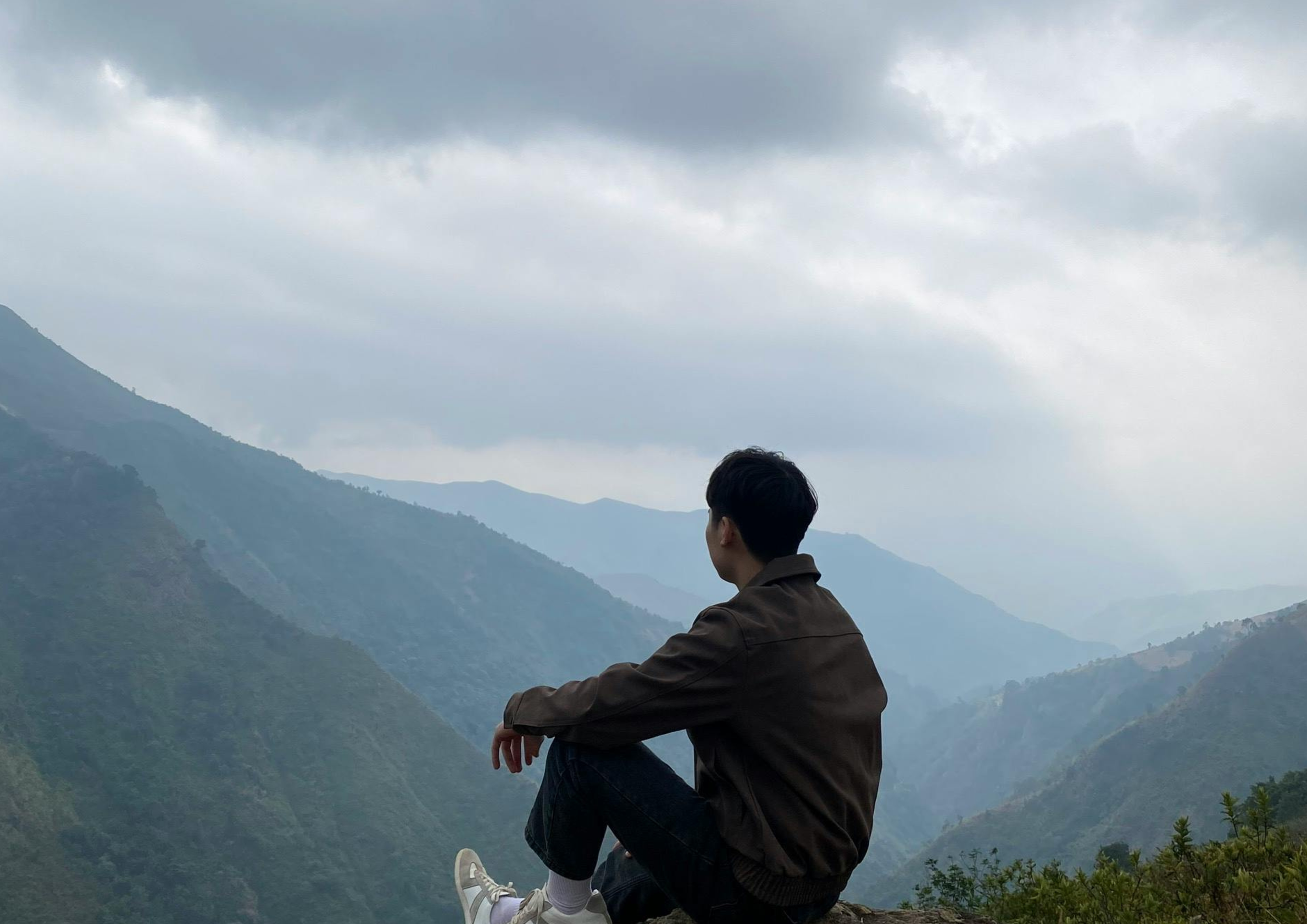 A person sitting on a mountaintop looking at a mountain range with cloudy sky.