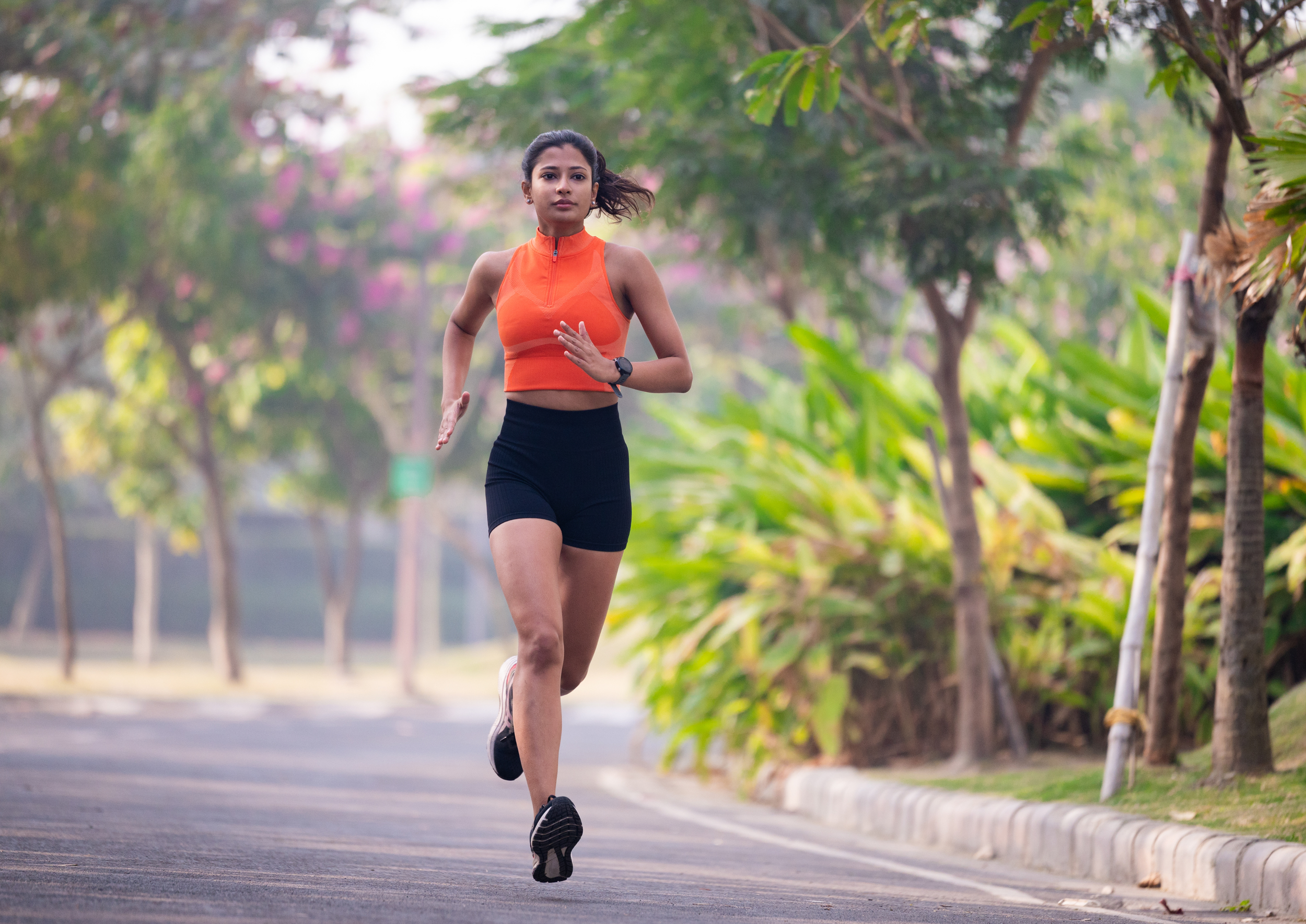 A woman running outdoors on a paved path surrounded by trees and greenery, wearing an orange sleeveless top and black shorts.