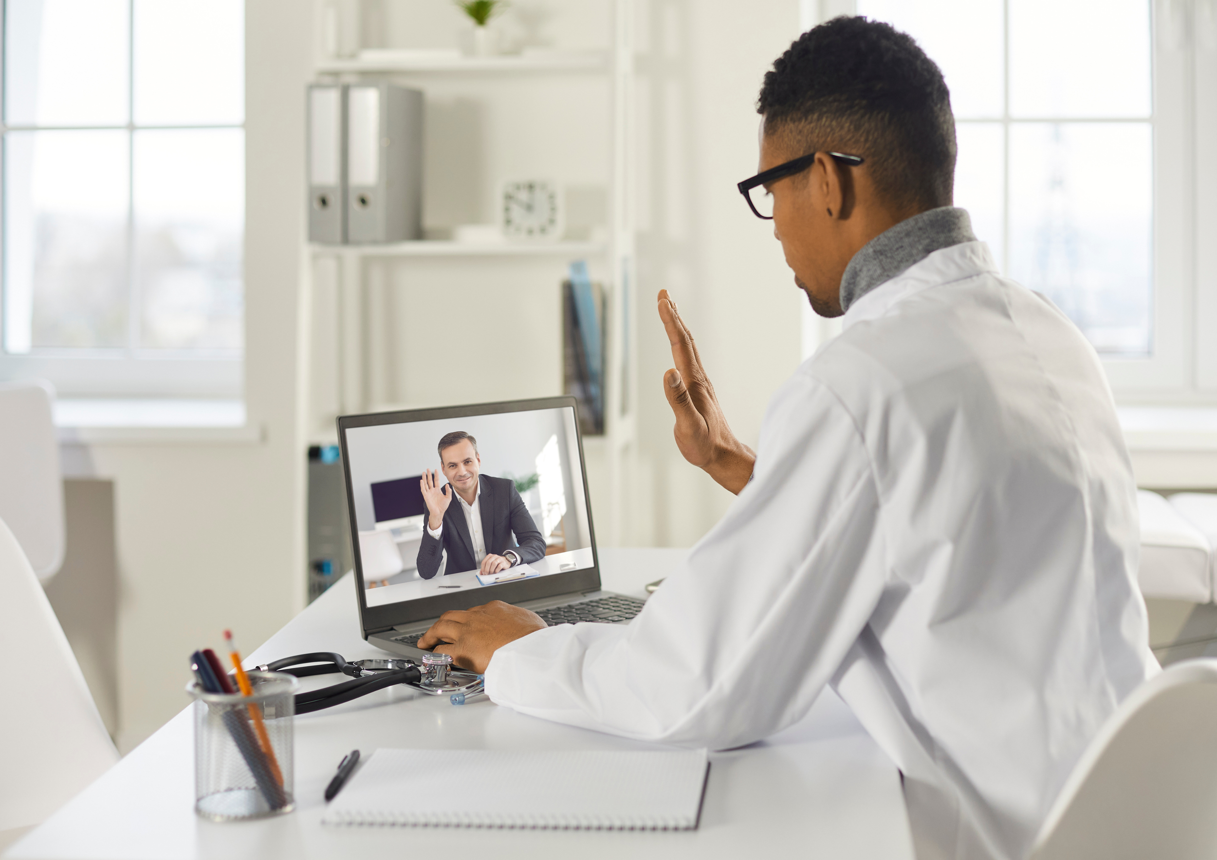 A doctor having a video call with a patient in an office. The doctor, wearing glasses and a white coat, sits at a desk with a laptop, raising his hand in a wave. The patient, visible on the laptop screen, is waving and wearing a dark suit.