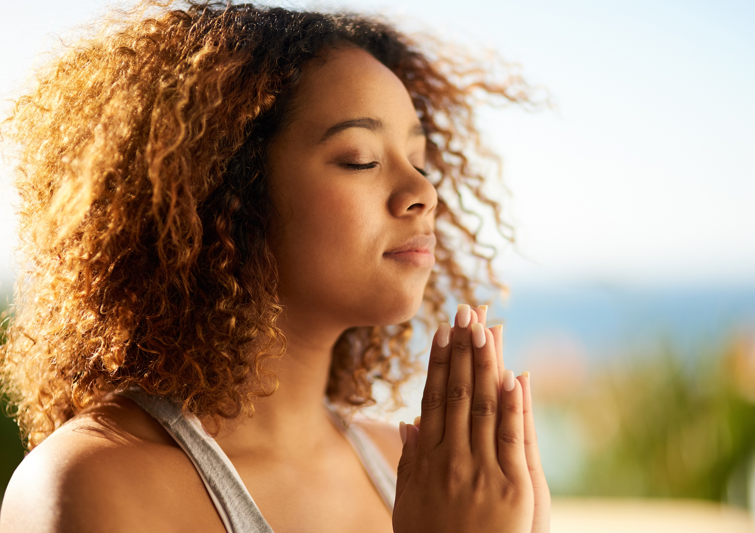 A woman with curly hair and closed eyes is practicing yoga or meditation outdoors with her hands pressed together in prayer position near her face.