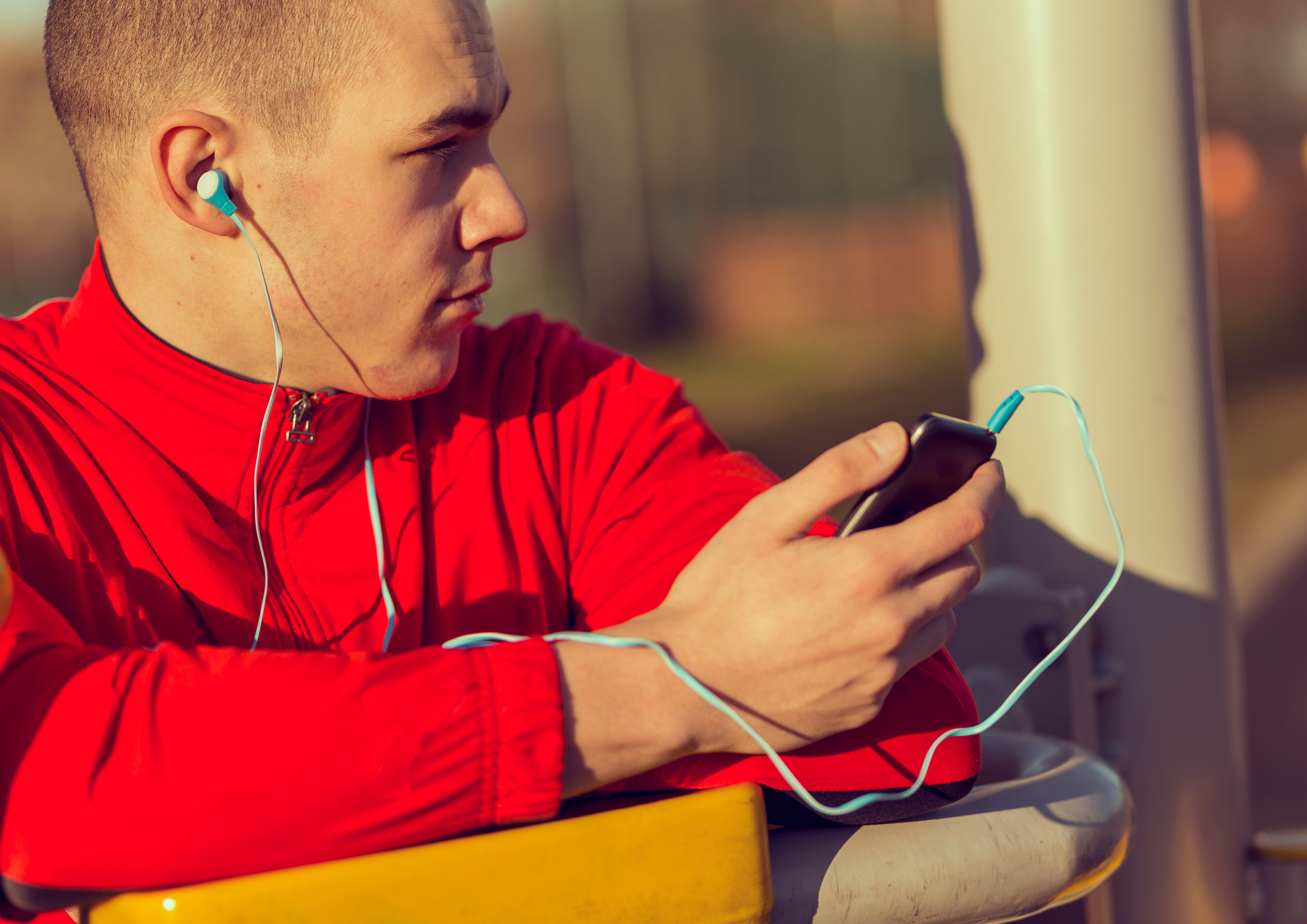 Young man in red jacket sitting on a bench outdoors, listening to music with earphones and looking at his phone.