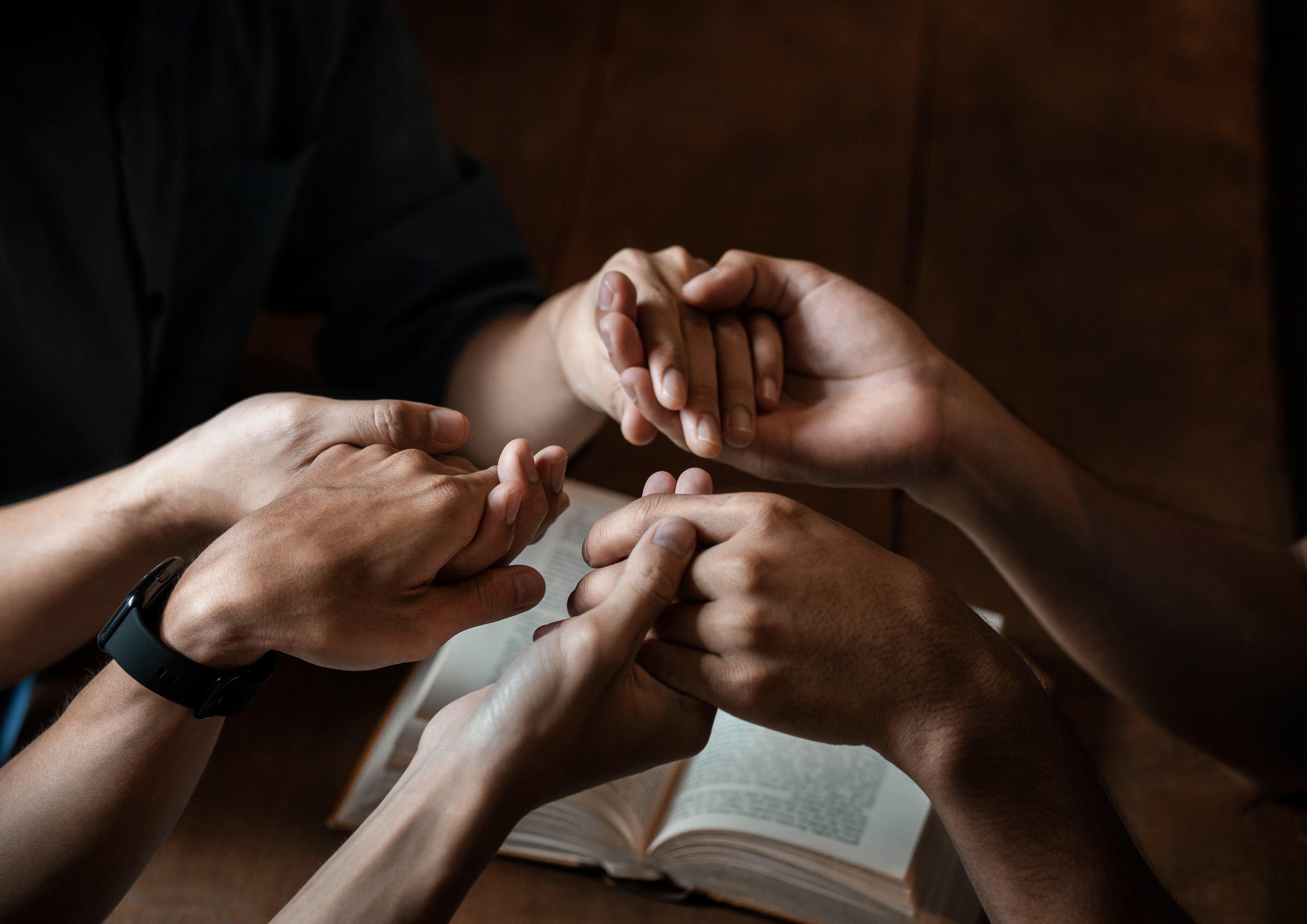 Multiple people holding hands in a circle above an open book on a wooden table.