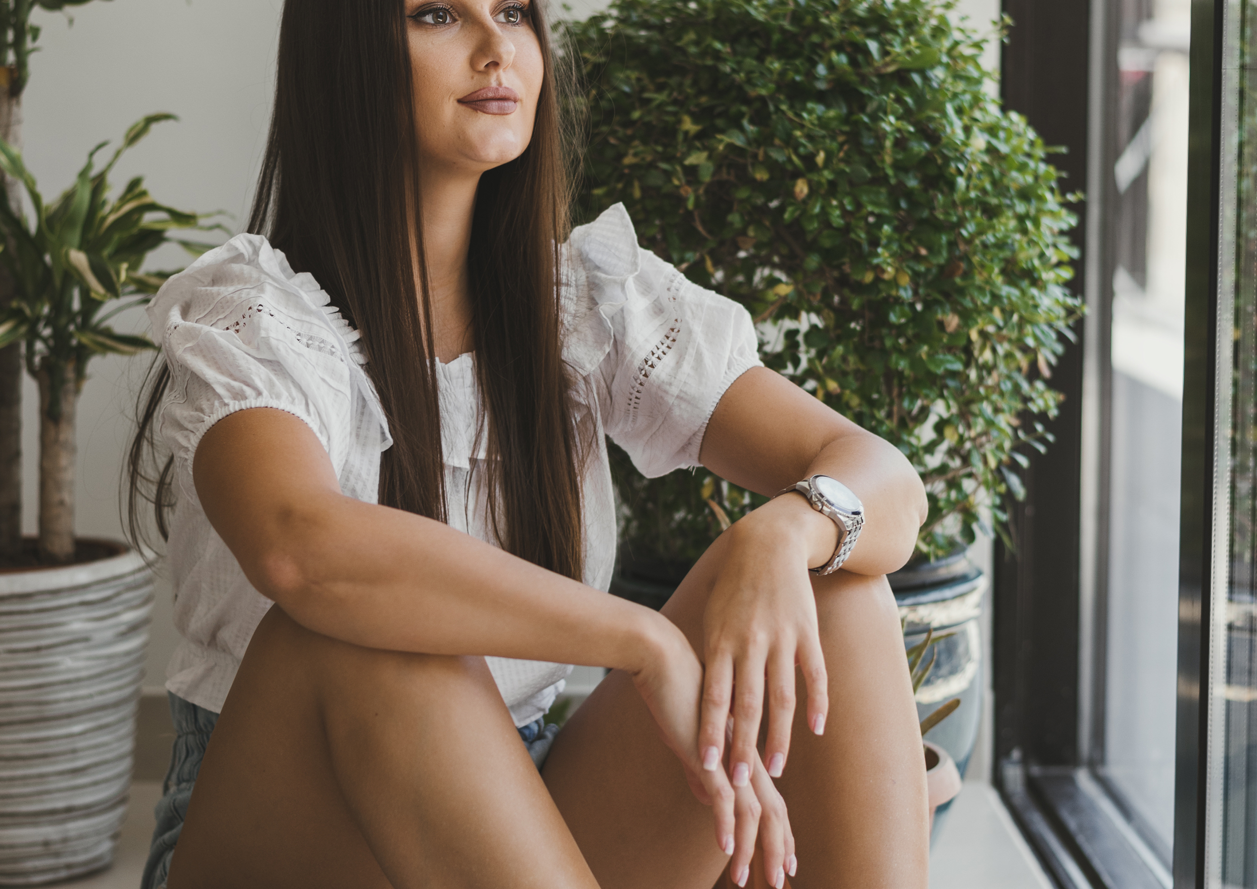 A young woman with long brown hair sitting indoors next to a large window, with green potted plants behind her. She is wearing a white short-sleeve blouse and denim shorts, and has a silver watch on her left wrist. She appears to be looking thoughtfully out the window.