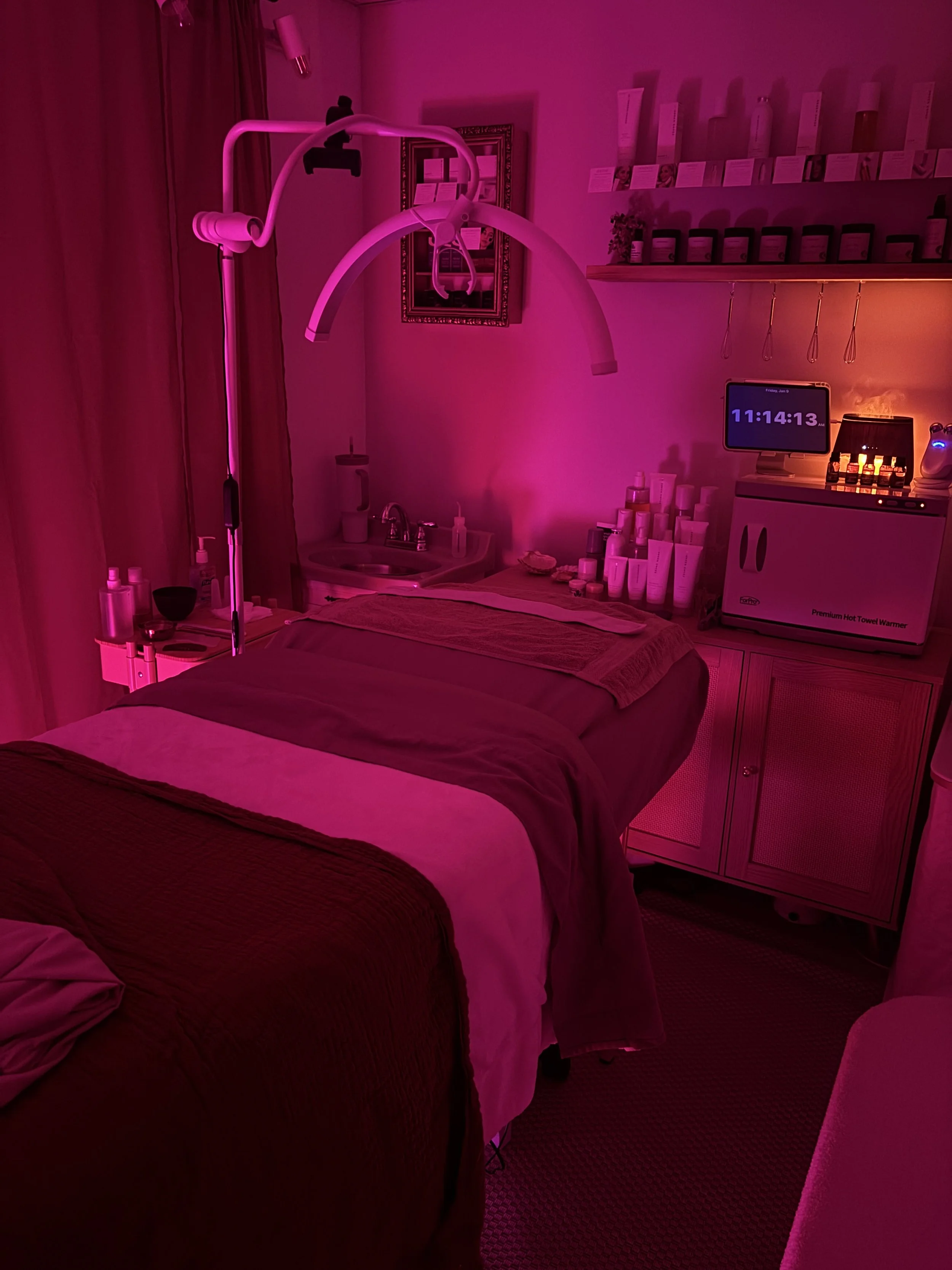 A treatment room with a massage table illuminated by pink lighting, featuring a variety of skincare products, a computer screen displaying the time, and medical equipment.