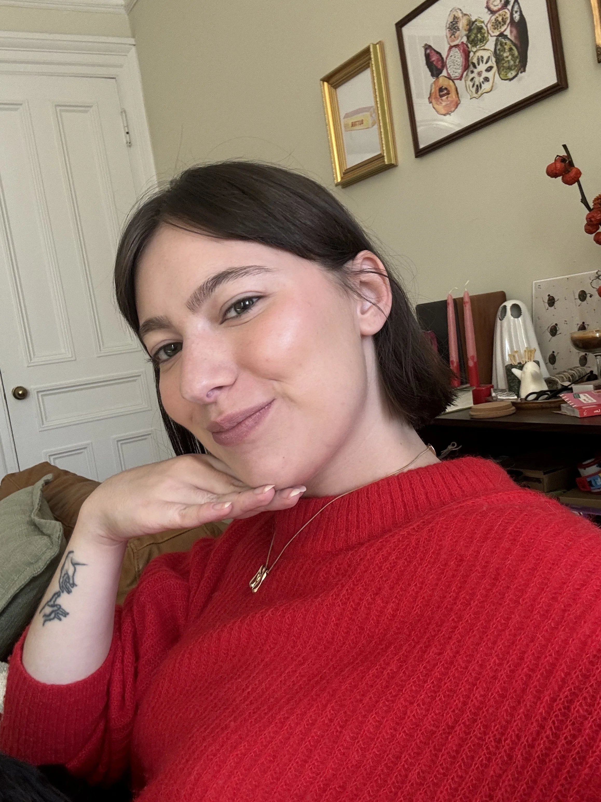A woman with short dark hair wearing a red sweater and a small gold necklace, smiling and resting her chin on her hand, in a room with framed art and decorations on the wall.
